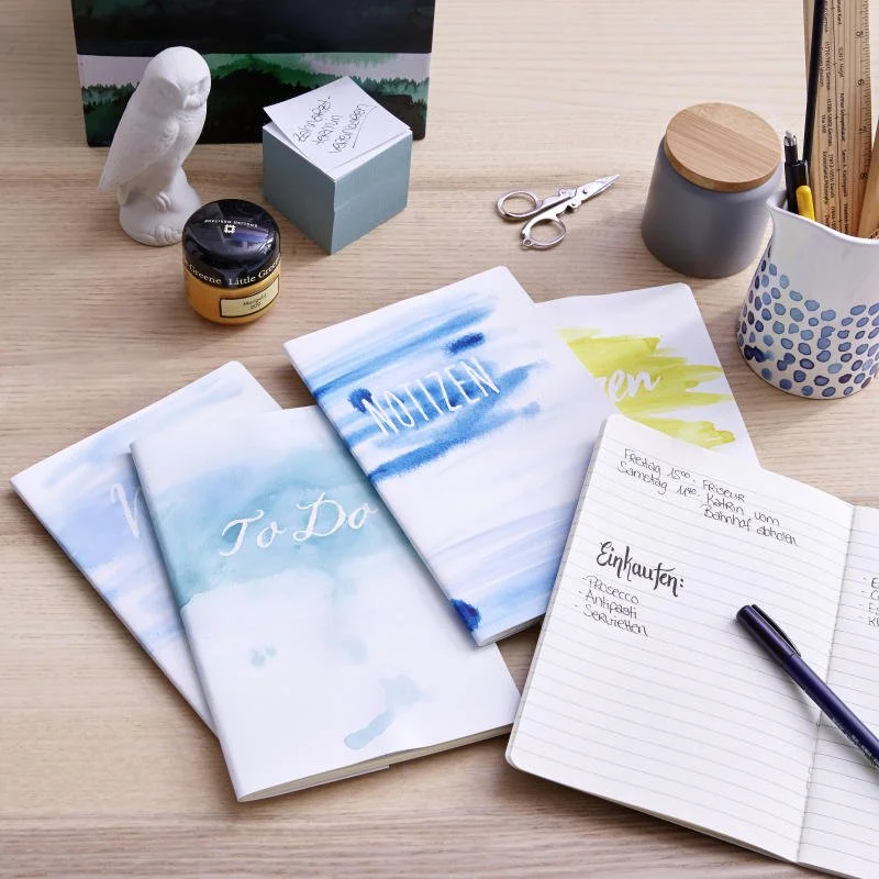 Ein Schreibtisch mit drei Notizbüchern, einem Notizbuch mit Einkaufsliste, einer schwarzen Kugelschreiber, einer Kerze, einer kleinen Statue eines Eulen, einer Schere, einem Behälter mit Holzdeckel, einem Ästchen mit Stift, einem Blumentopf mit blauer Punkte-Dekoration und einem Bild im Hintergrund.