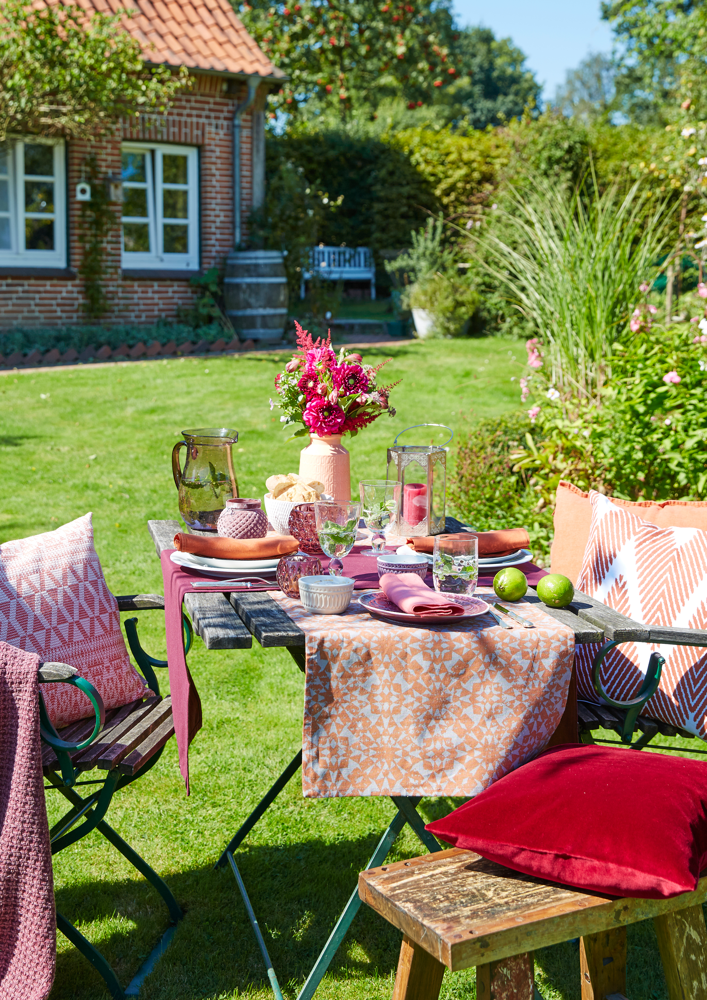 Ein Gartentisch ist für ein Essen im Freien gedeckt, mit einer bunten Tischdecke, Geschirr, Gläsern, einer Vase mit pink-rote Blumen und zwei grünen Limetten. Es gibt bunte Kissen auf Stühlen und Bänken, umgeben von einem gepflegten Garten mit grünem Rasen, Büschen und einem Haus im Hintergrund.