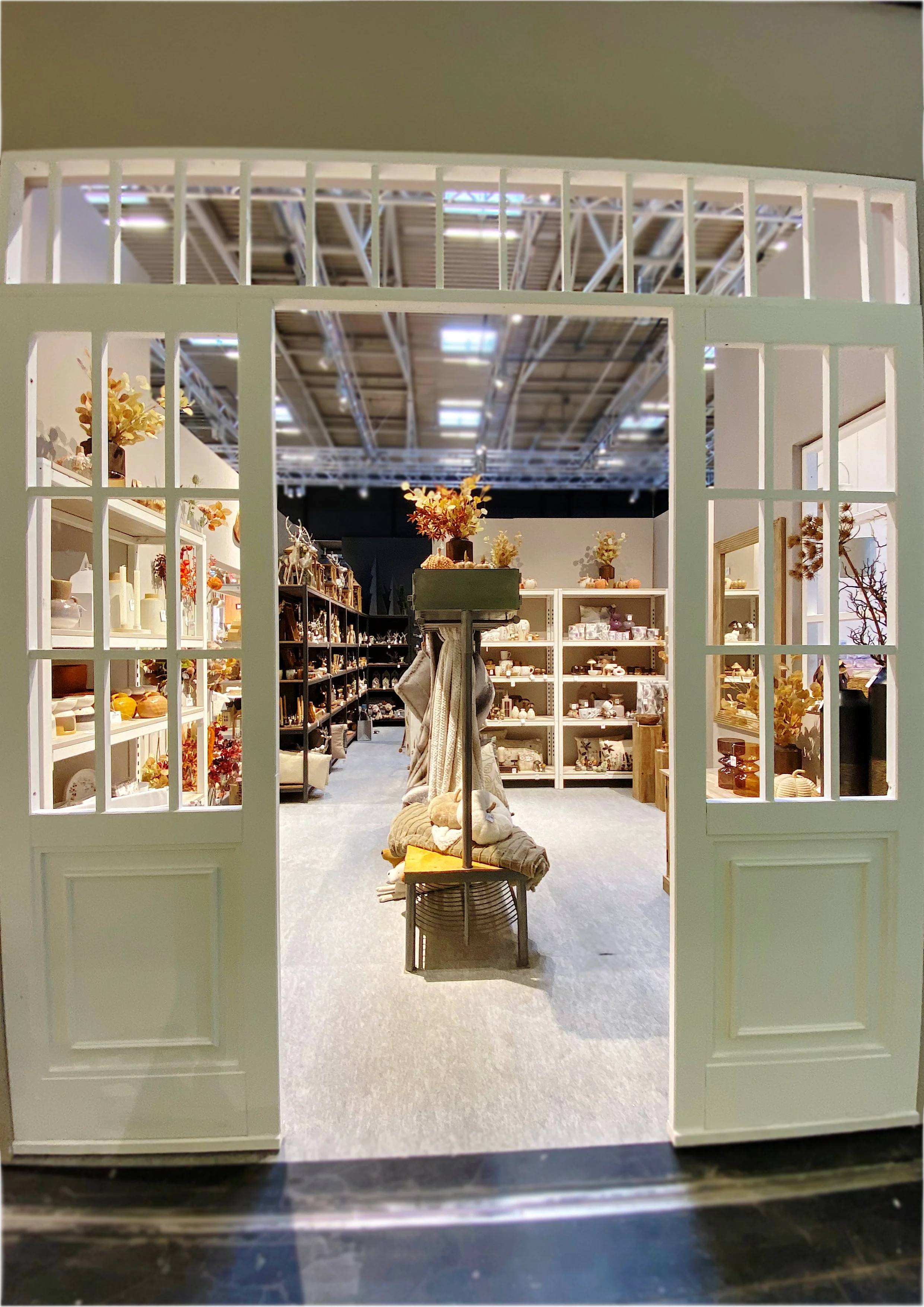 Blick durch die offene weiße-Tür in einen Laden mit Regalen, die mit Dekorationsartikeln, Vasen und Pflanzen gefüllt sind. Im Zentrum steht eine Stuhlbank mit dekorativen Gegenständen.