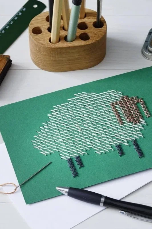 Bastelarbeit auf grünem Papier, das einen Schaf mit Filzstiftfarben zeigt, mit einem Holzuntergrund, einem Dreiecknadel, einem Kugelschreiber, einem Holzstift und einem Holzorganisator für Stifte im Hintergrund.