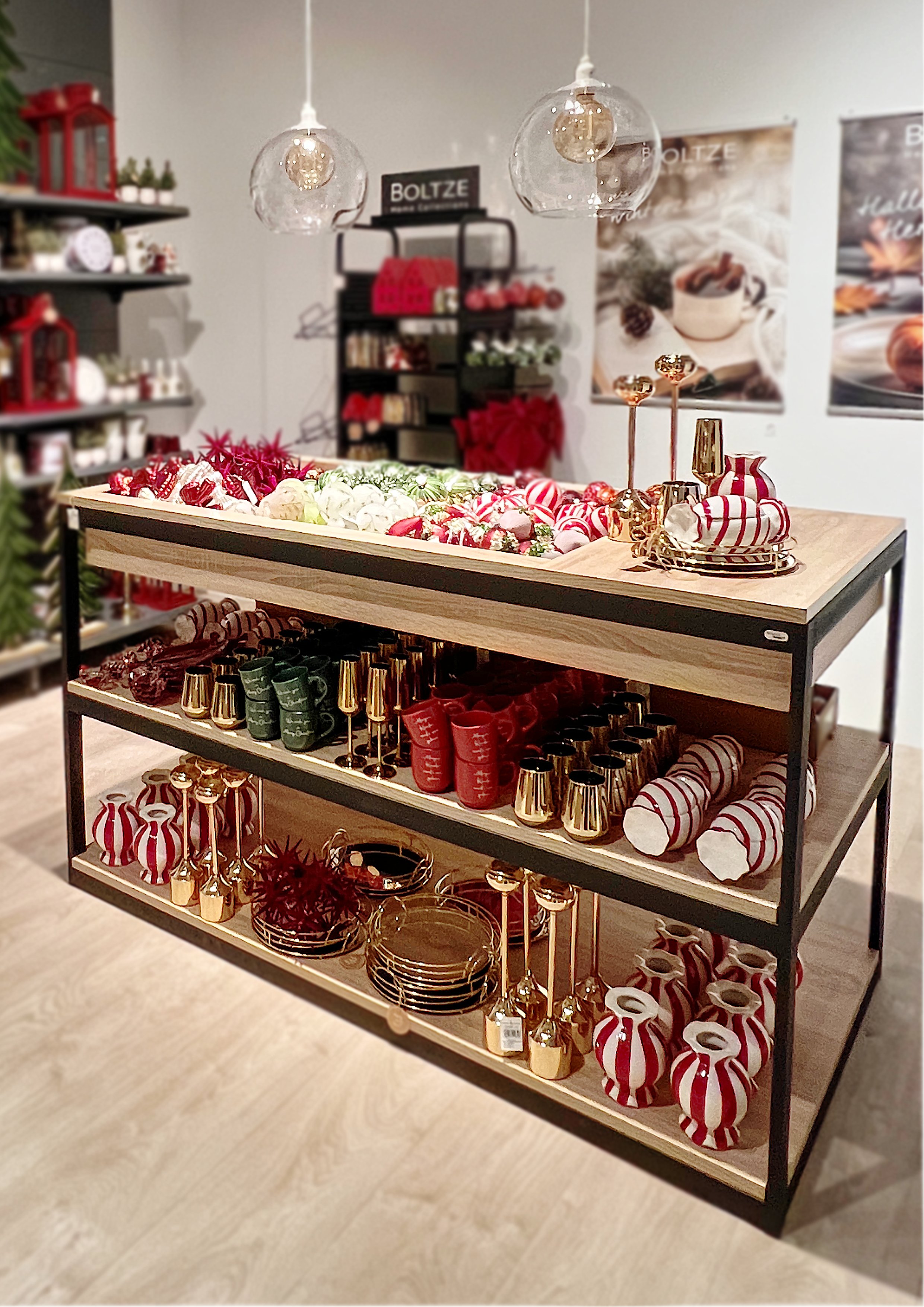 Dekorative PoS-Präsentation von Weihnachtsartikeln auf einem dreistöckigen Tisch, darunter Vasen, Kerzen, Geschirr und Dekorationen in Rot, Weiß, Grün und Gold.