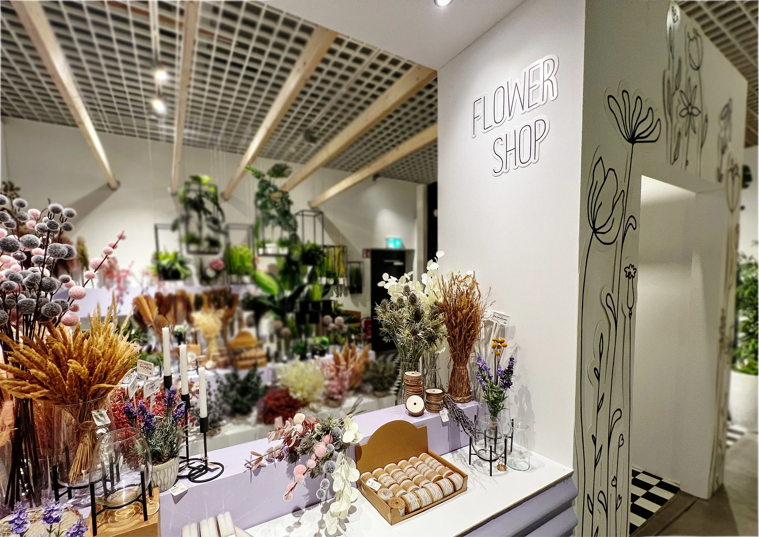 Blumenladen mit Deko und Blumenarrangements in einem Laden namens Flower Shop, weißer Wand mit gestalterischer Blumenzeichnung, Tische mit verschiedenen Blumen und Dekoartikeln, Holzdecke.