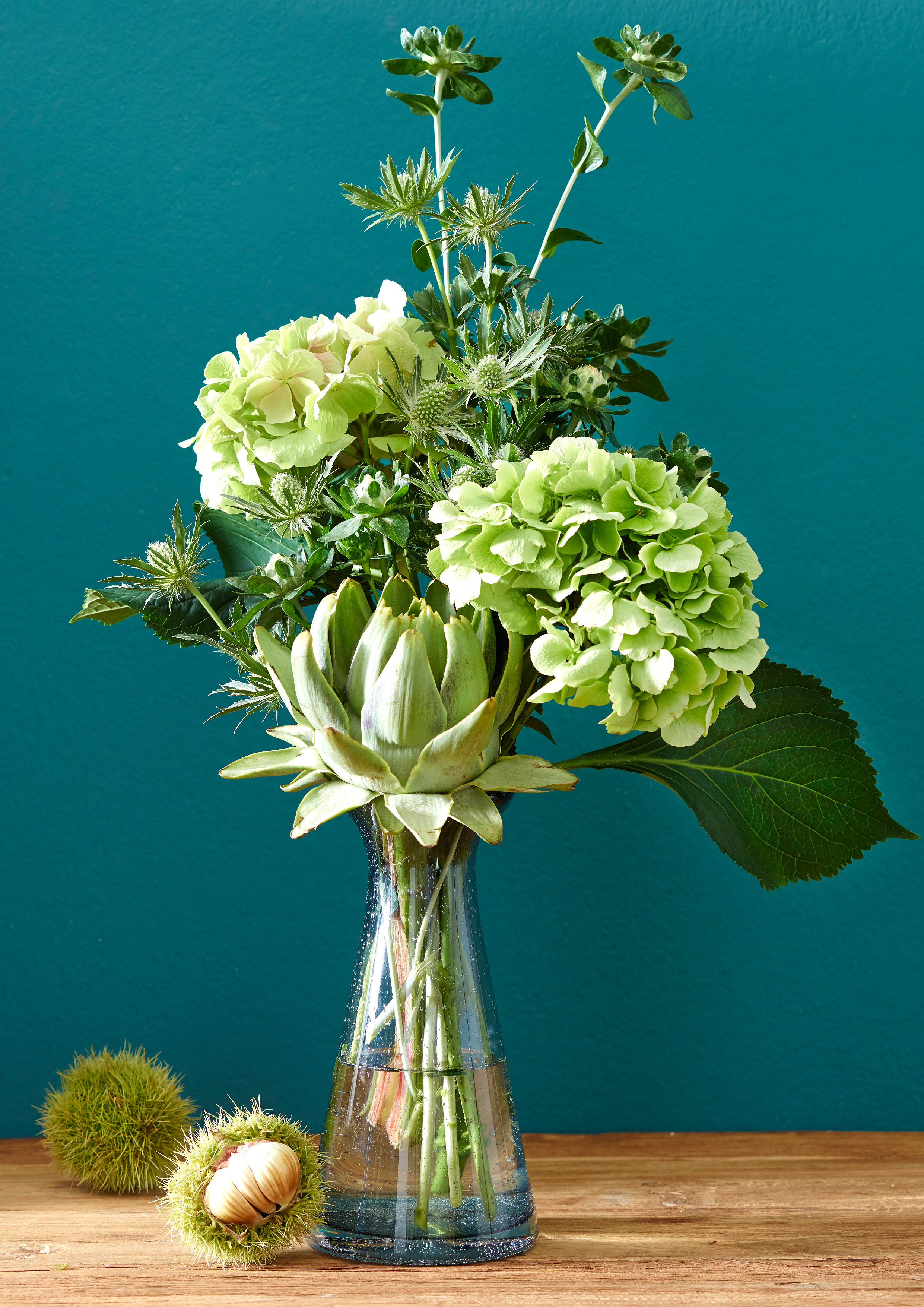 Blumenstrauß mit hellgrünen Hortensien, Artischocke und verschiedenen grünen Blättern in einer Glasvase auf einem Holztisch vor einer grünen Wand.