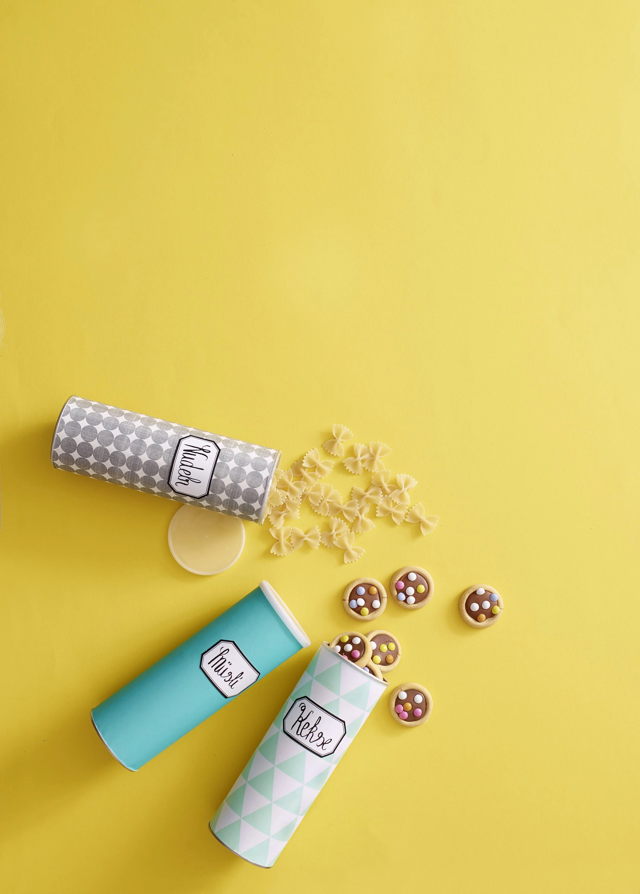 Drei bunte Keksdosen mit Namen, eine offene Dose mit Farfalle-Nudeln, eine geschlossene Dose mit Mürbiteig-Keksen, und einige Kekse mit bunten Zuckerperlen auf gelbem Hintergrund.