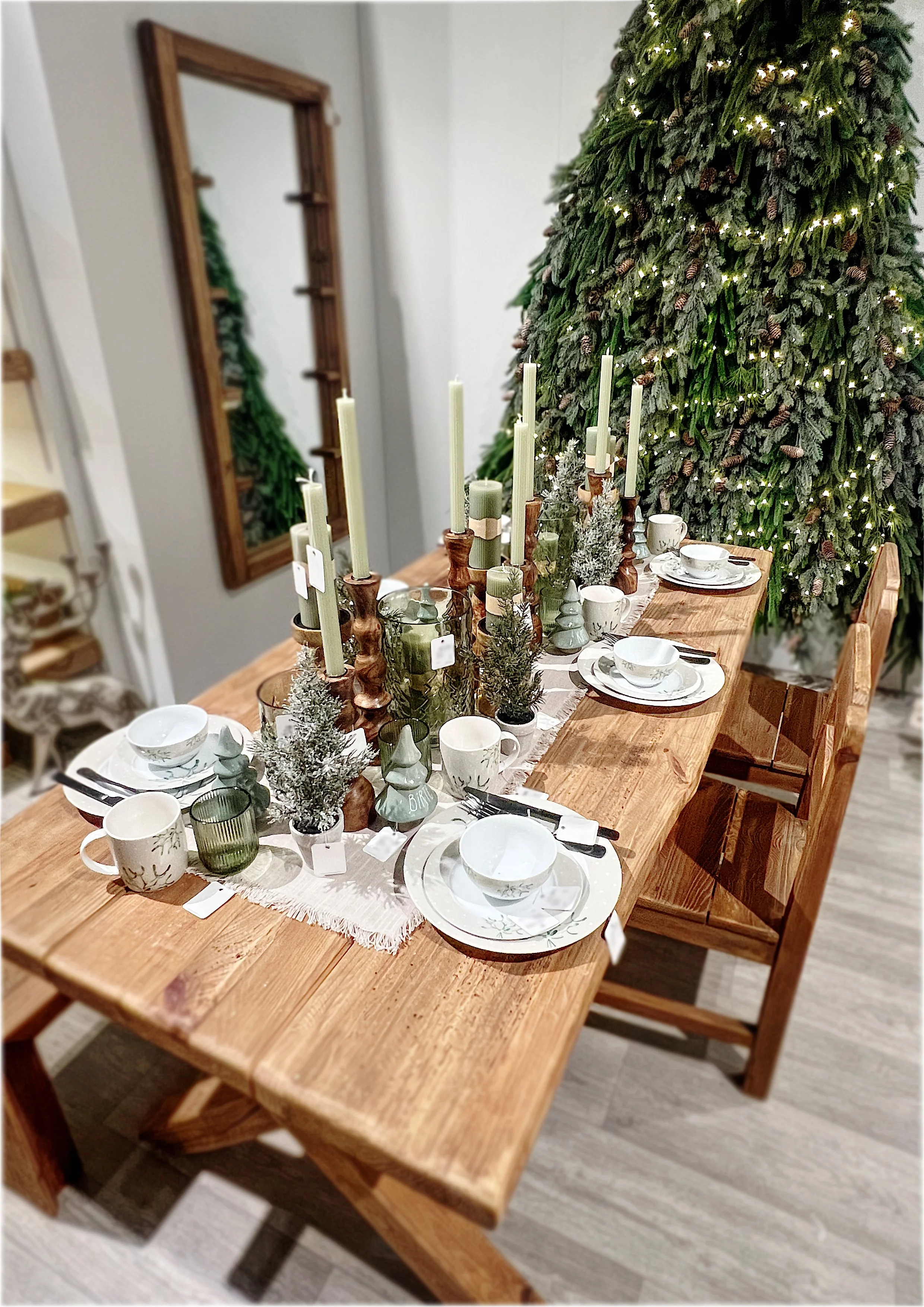 Ein festlich gedeckter Esstisch mit Weihnachtsdeko, umliegender Tanne, Kerzen und Geschirr.