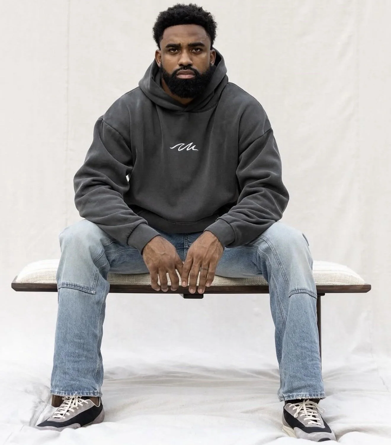 A man with a beard and short curly hair sits on a bench against a plain white background, wearing a dark hoodie, light blue jeans, and sneakers, looking directly at the camera with a serious expression.