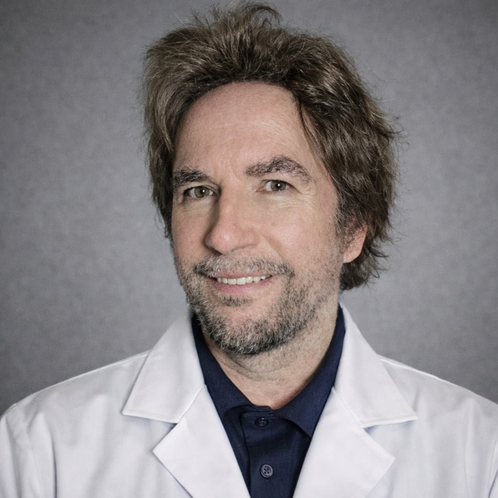 Portrait of a man with brown, slightly tousled hair, light-colored eyes, and a beard, wearing a white lab coat over a dark blue shirt, against a gray background.
