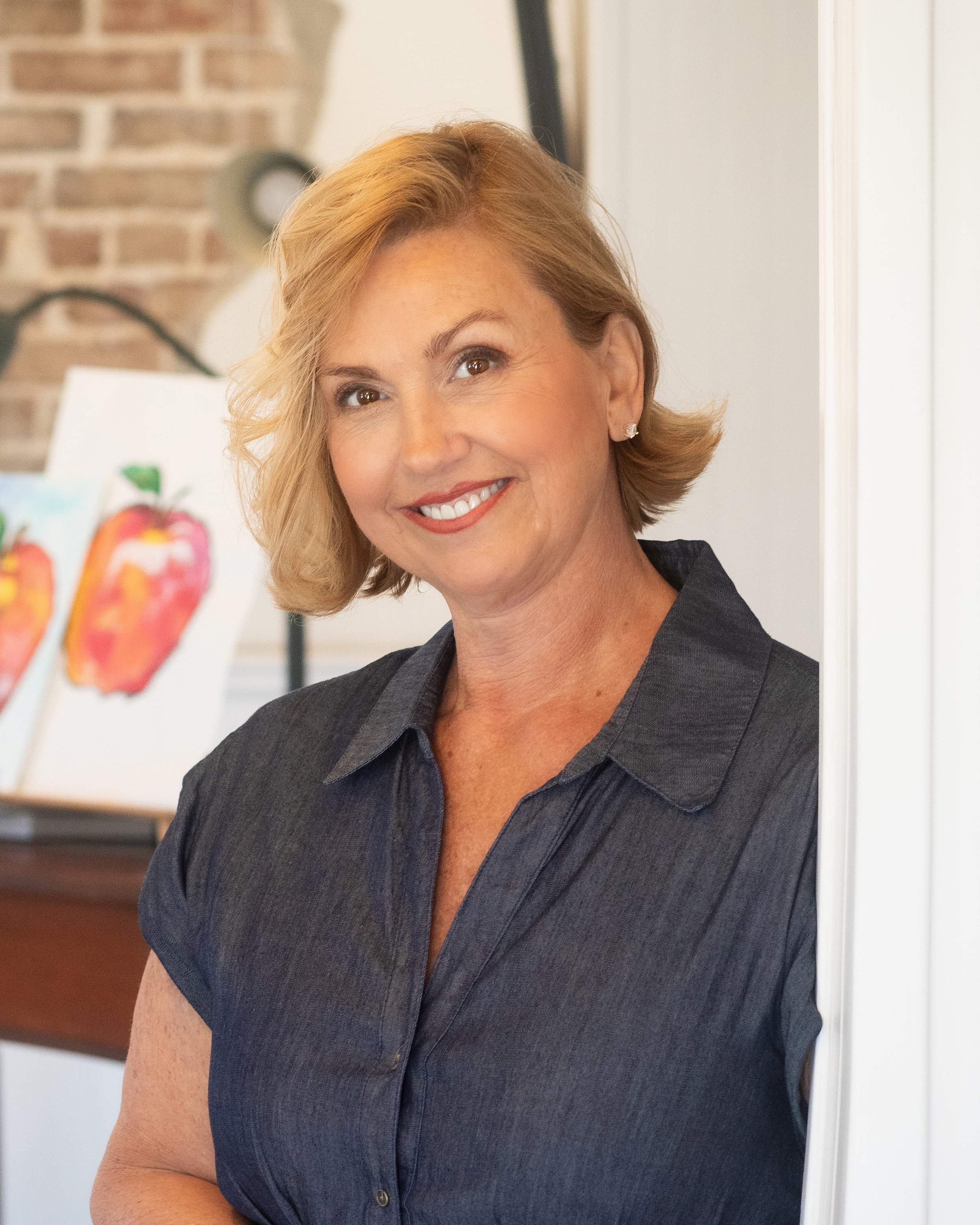 Close-up of a smiling middle-aged woman with short blonde hair, wearing a dark blazer, standing indoors with a brick wall and artwork in the background.