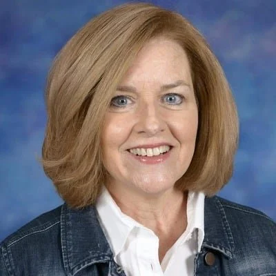 A woman with shoulder-length blonde hair, blue eyes, smiling, wearing a white shirt and denim jacket against a blue textured background.