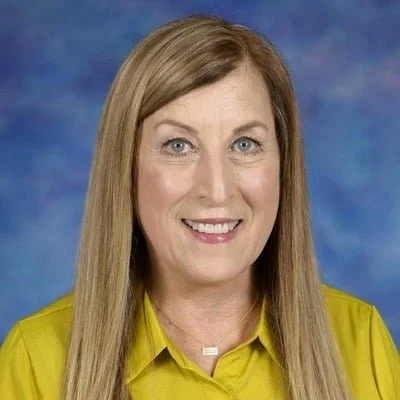 A woman with long blonde hair, wearing a yellow blouse and a small necklace, smiling at the camera against a blue backdrop.