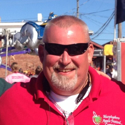 A smiling man wearing sunglasses and a red shirt with an apple logo at the Mississippi Apple Festival.
