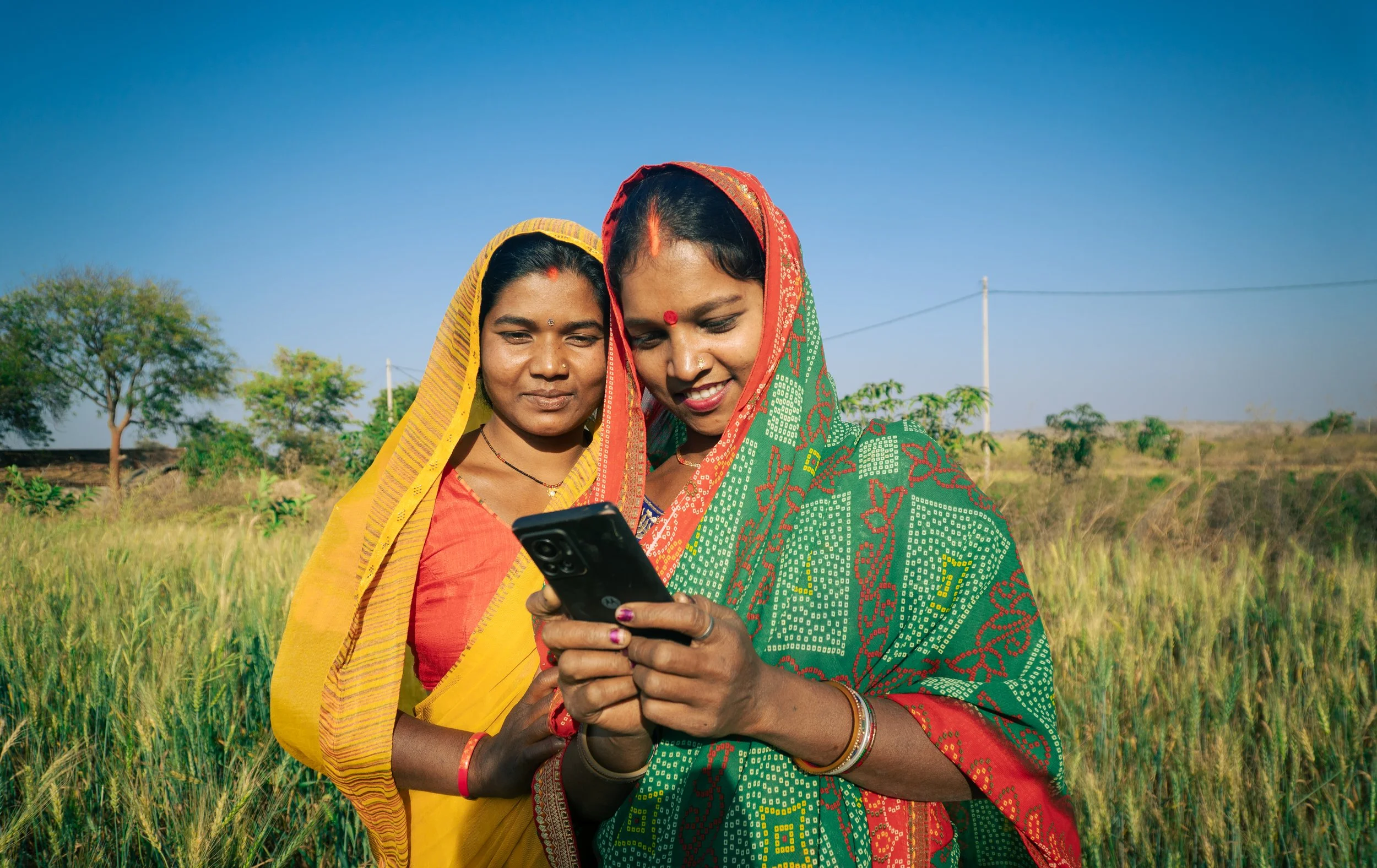 Two women in traditional Indian sarees standing in a field, looking at a smartphone together, with trees and a blue sky in the background.