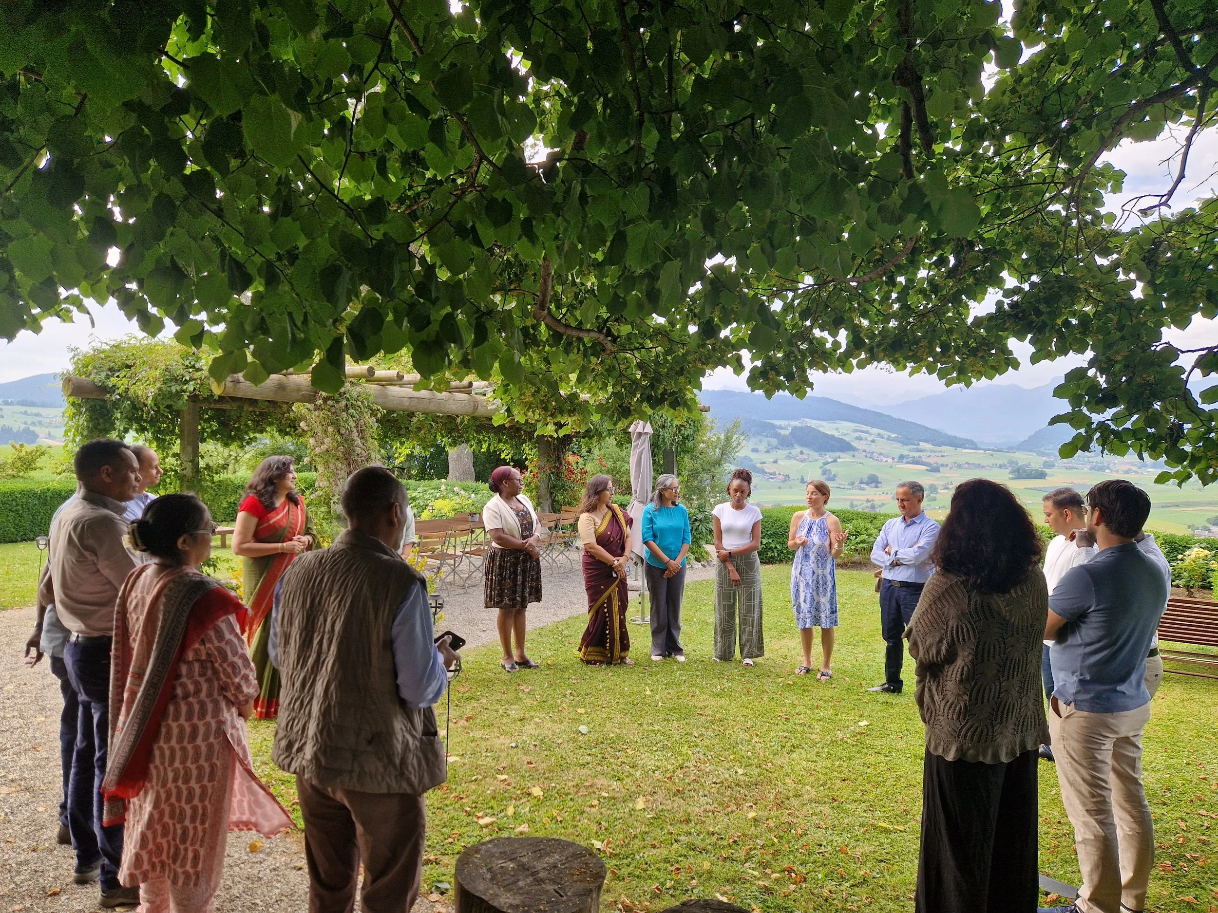 A group of diverse people standing in a circle outdoors under a large leafy tree, with a scenic view of hills and fields in the background.