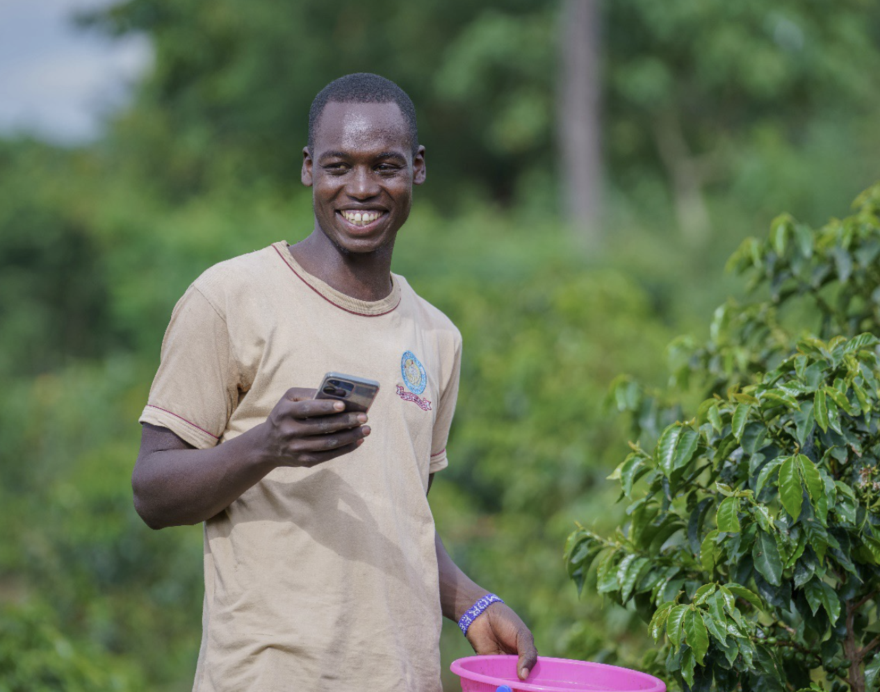 A young man with dark skin, smiling and holding a smartphone in his right hand, standing outdoors in front of green vegetation, with a pink bucket in his left hand.