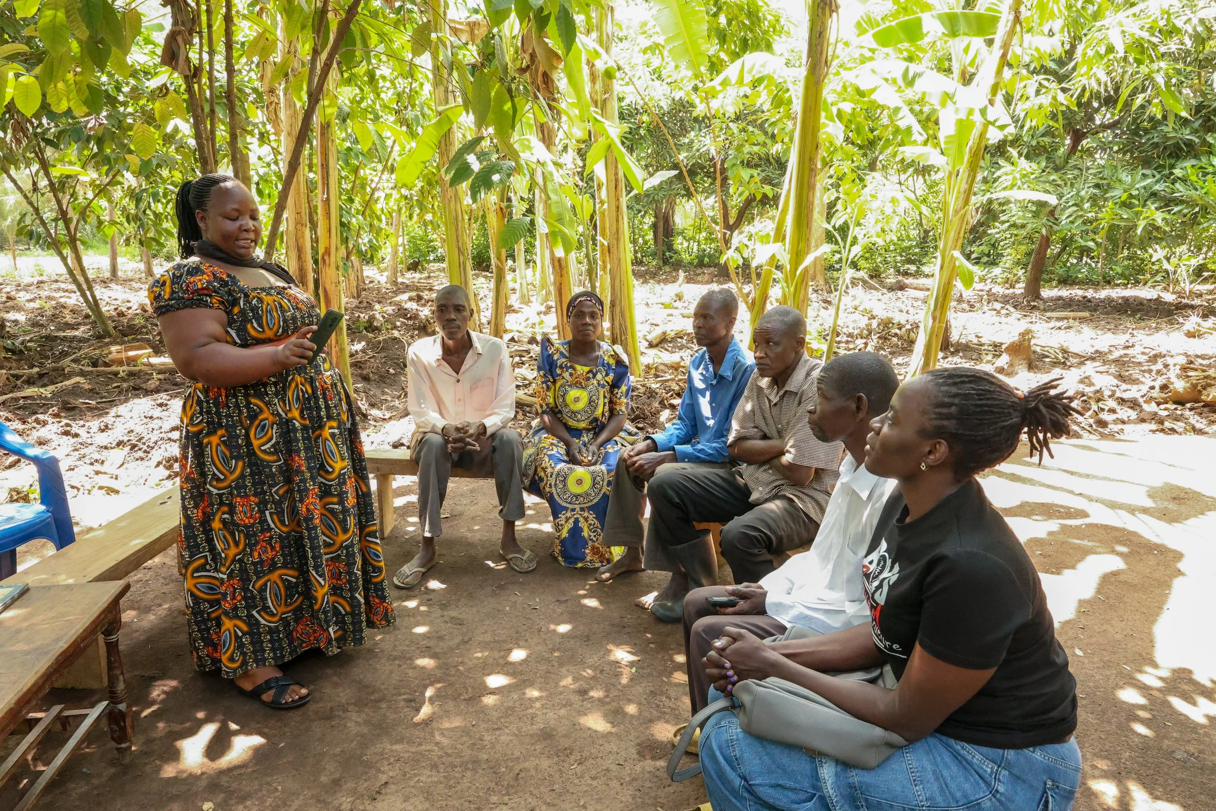 A woman standing and speaking to a group of people sitting on benches outdoors under trees.