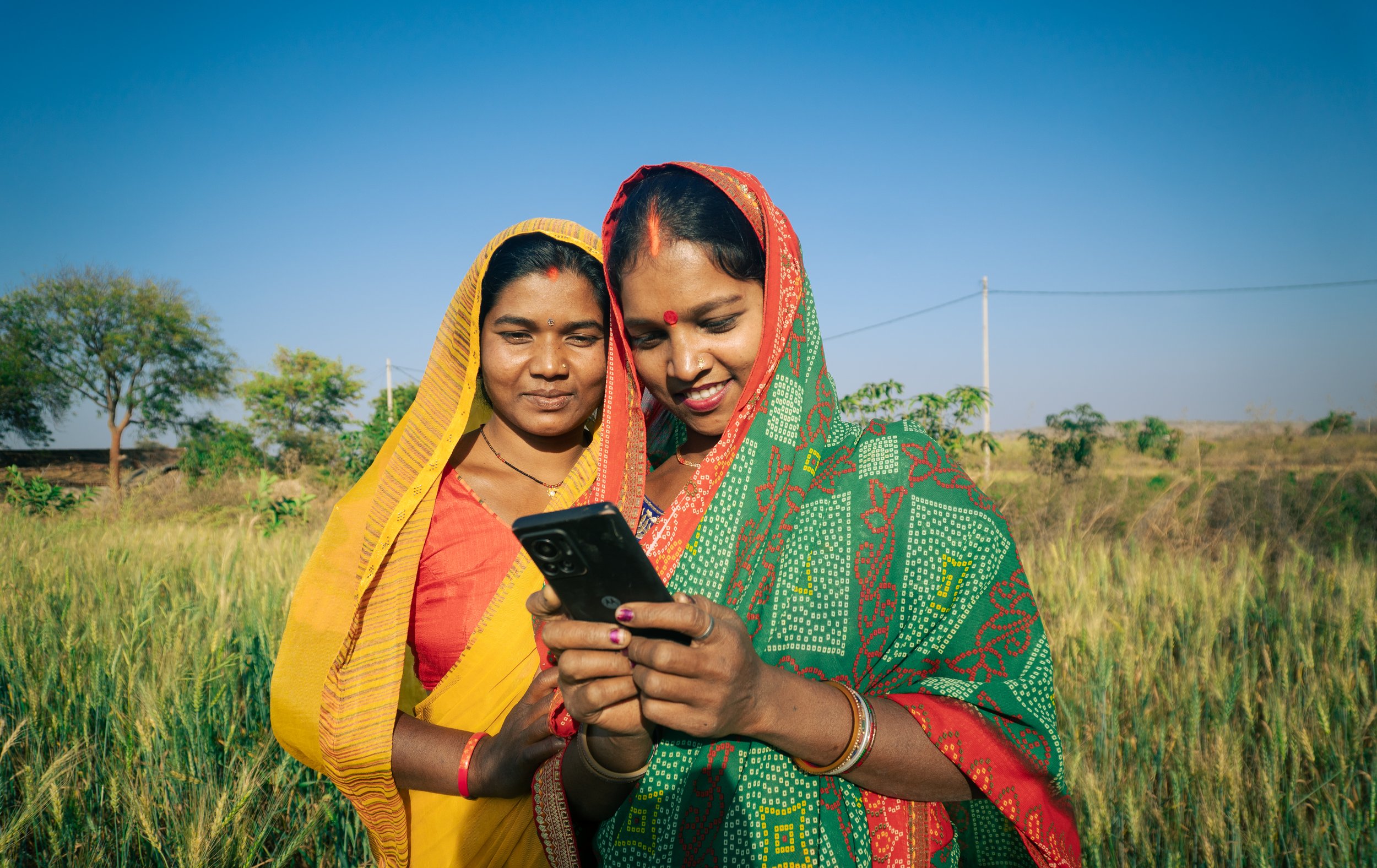 Two Indian women in colorful sarees looking at a smartphone in a field of tall grass with trees and a clear blue sky in the background.