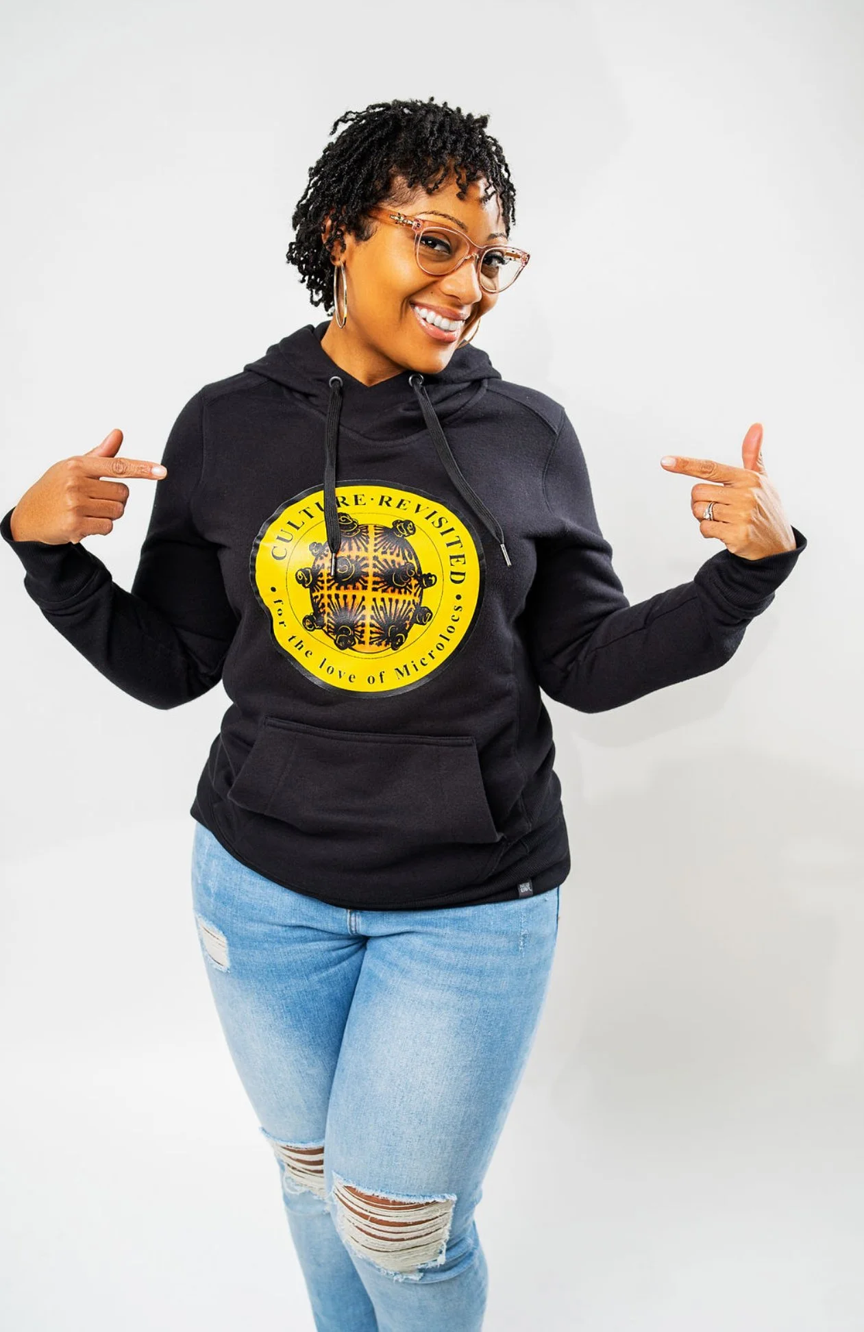 A woman with glasses and hoop earrings smiling and pointing to her black hoodie with a yellow circular logo that reads 'Culture Revisited' and 'For the love of MicroLocs', dressed in light blue ripped jeans against a plain white background.