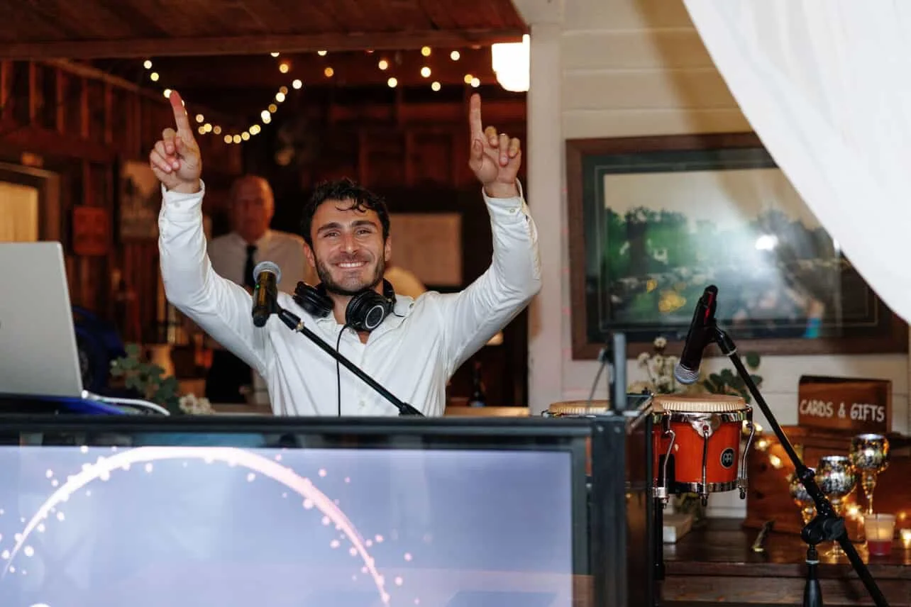 A smiling DJ with headphones around his neck at a party, raising both hands with index fingers pointing upwards, in a warmly lit room with string lights, a microphone, a DJ setup including a laptop, and a large monitor in the background.