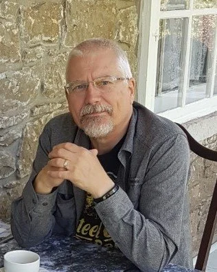 A photo of the author, Glenn Kerridge, sitting at a table with his hands clasped, in front of a stone wall and window, with a white cup on the table.