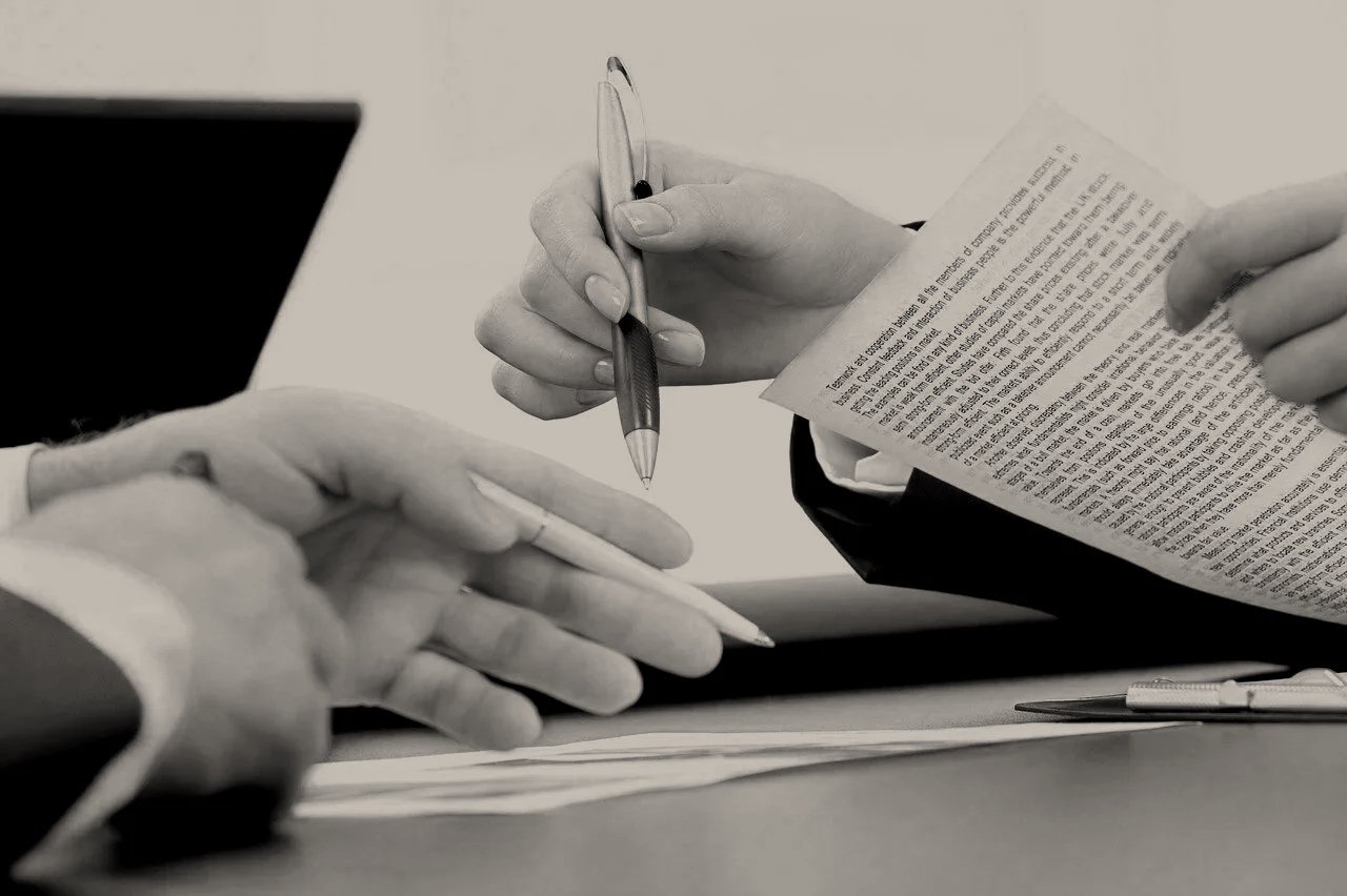 Manos en reunión de negocios, una persona entrega un documento a otra mientras otra más toma notas con una pluma en un escritorio