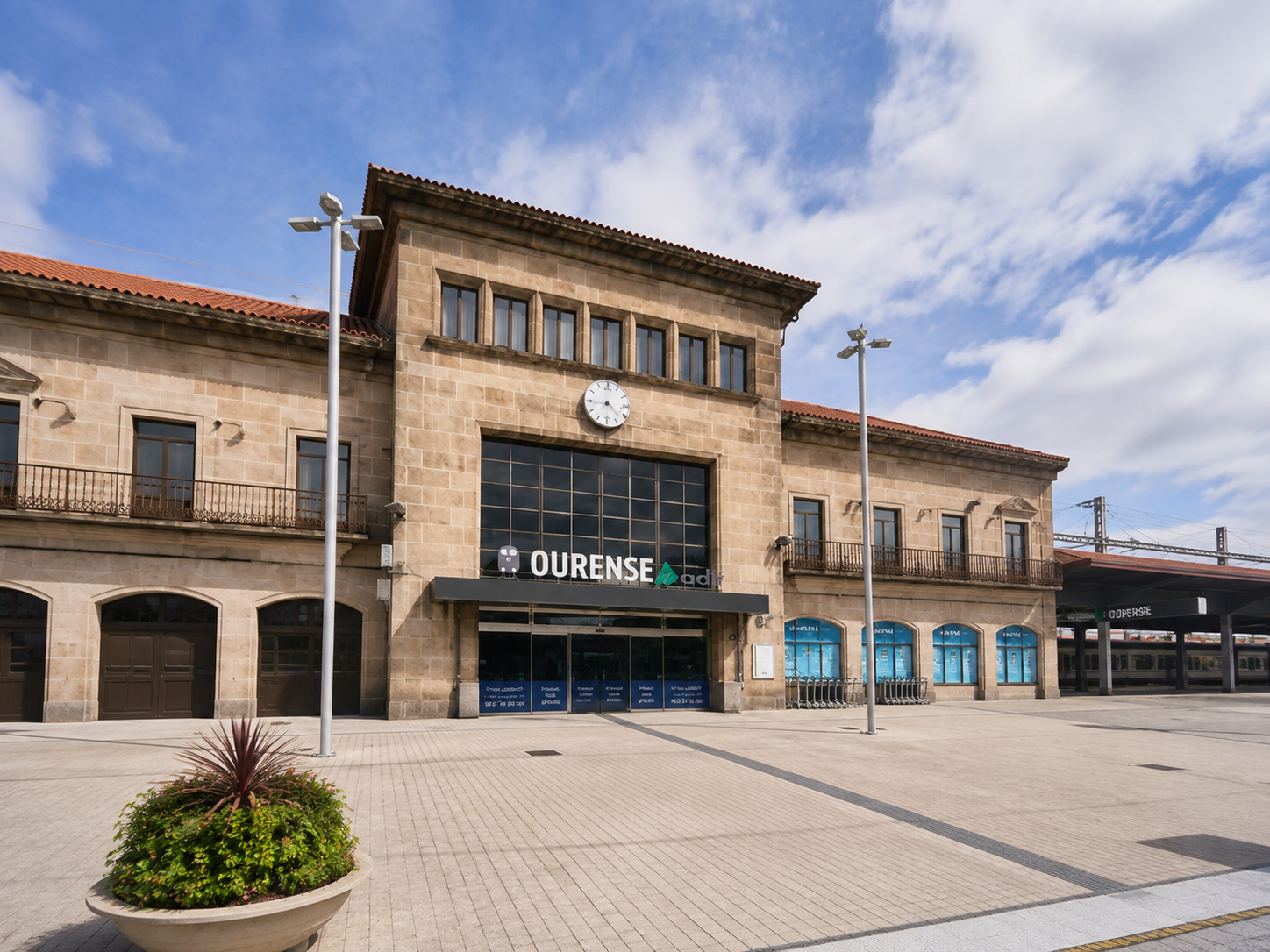 Estación de tren en Ourense con fachada de piedra, reloj, señal de adiós y bancos en la parte exterior, bajo cielo parcialmente nublado.