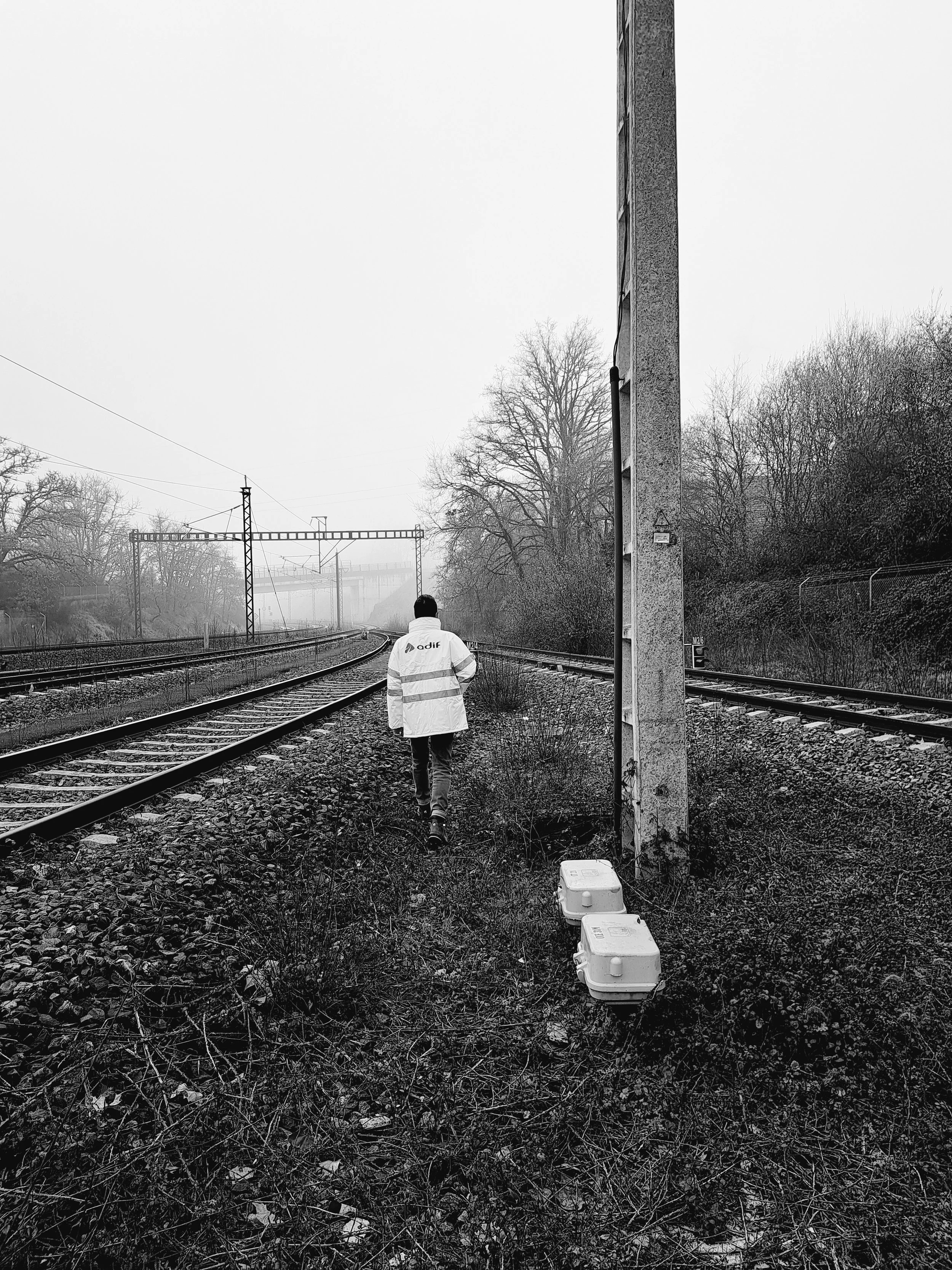 Persona caminando al lado de las vías de tren en una tarde nublada, con árboles sin hojas y dos cajas en el suelo.