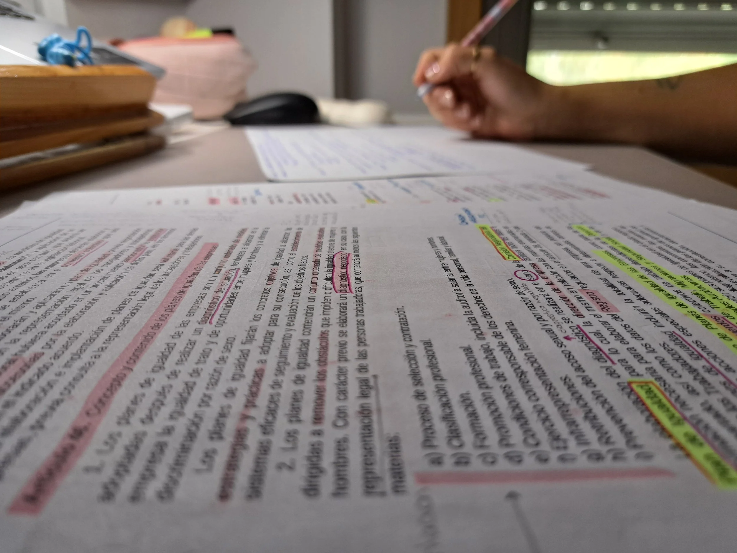 Páginas impresas con resaltadores y anotaciones, en primer plano, en un escritorio. En el fondo, una mano escribiendo y objetos de oficina.
