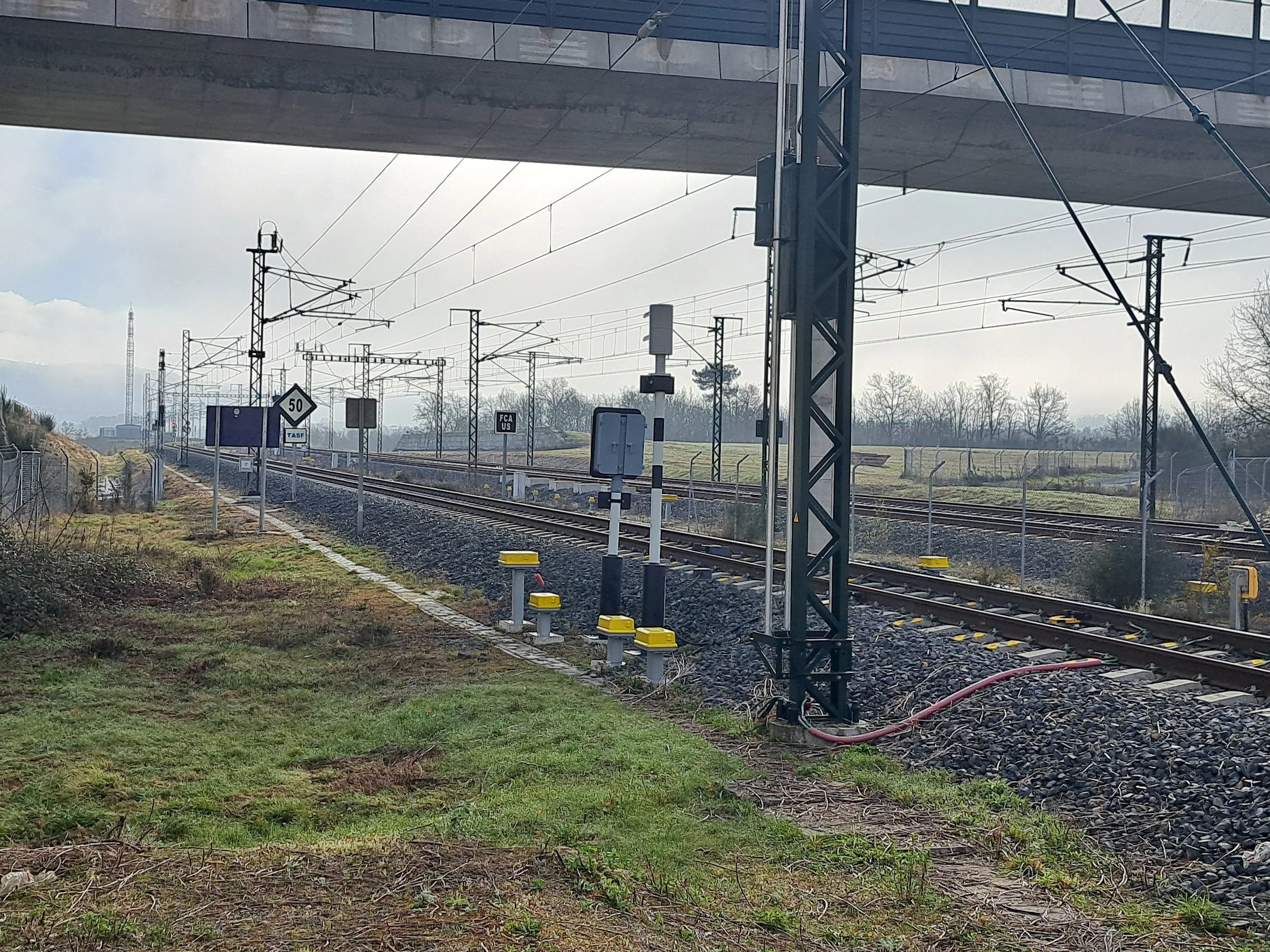 Vías de ferrocarril vistas desde debajo de un puente, con señales y cables eléctricos en un entorno rural.