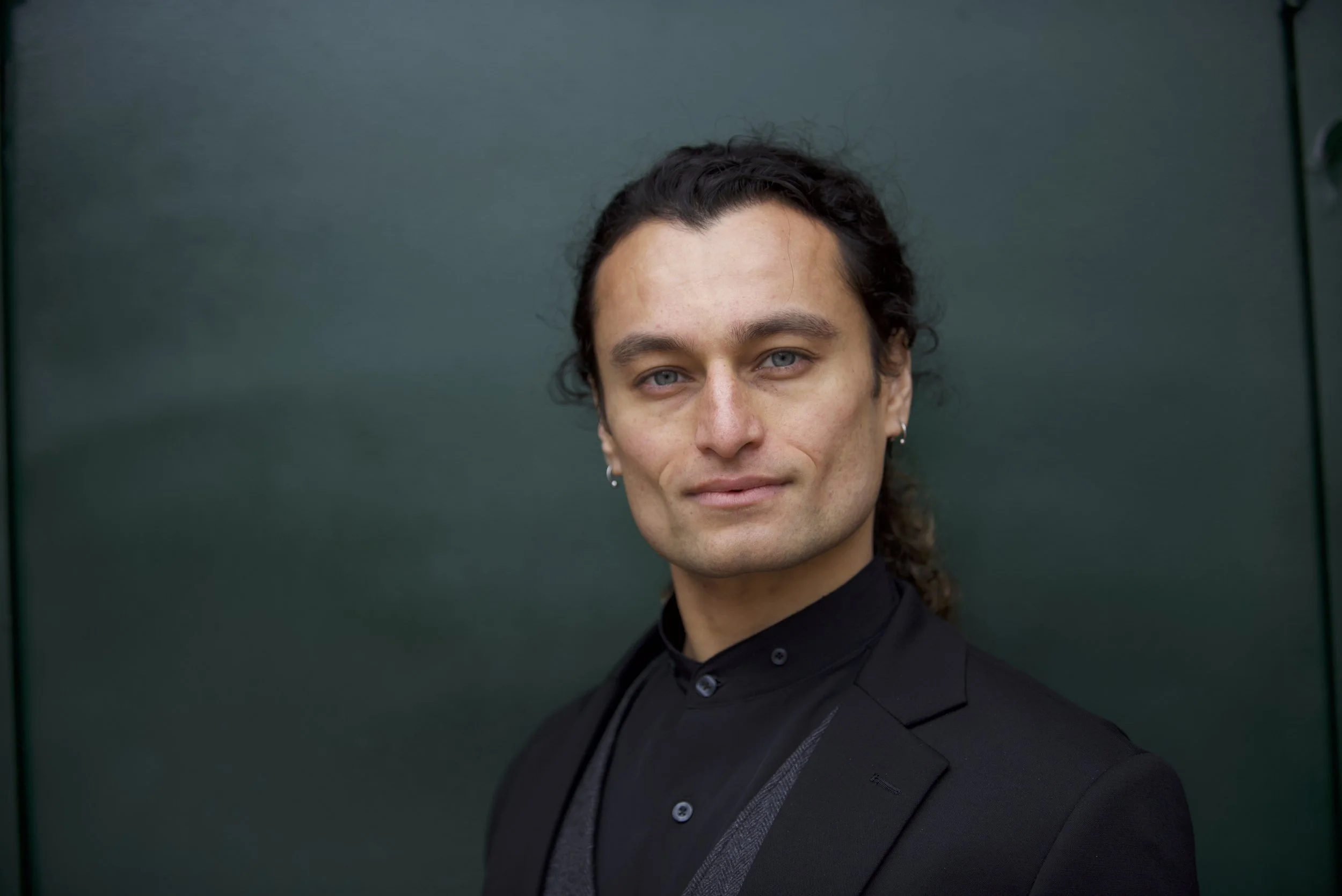 A man with long dark hair, light skin, and blue eyes, wearing earrings, a black shirt, and a black blazer, standing in front of a dark green background.