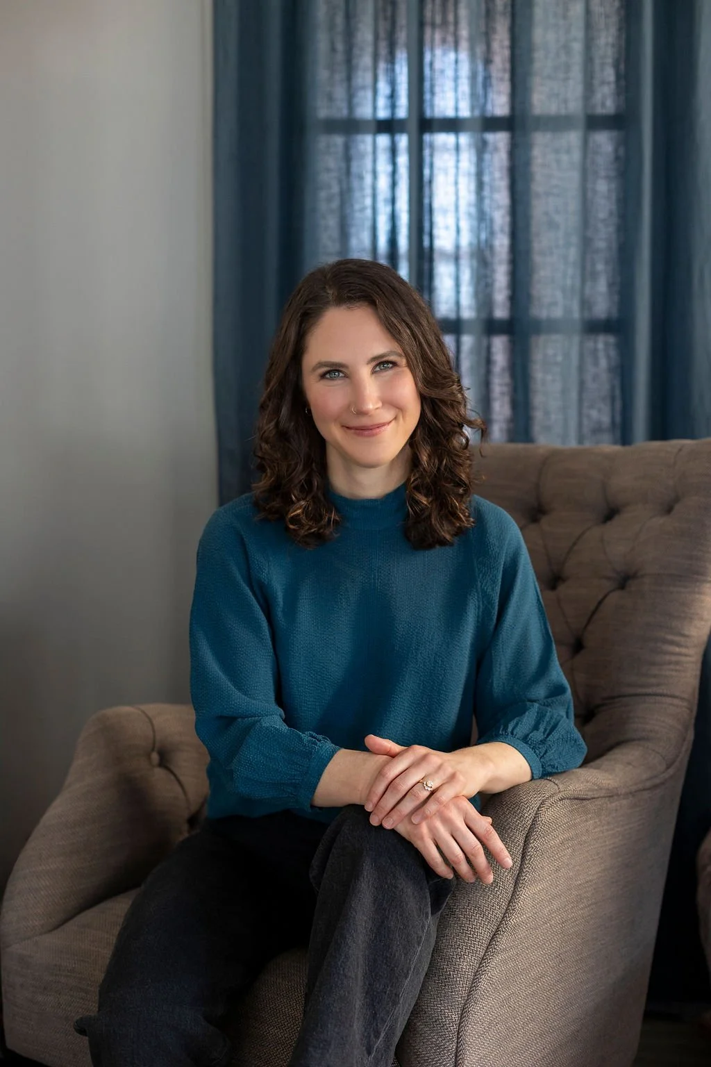 A woman with brown, curly hair and blue eyes smiling, sitting on a beige armchair in a room with blue curtains and a window in the background.