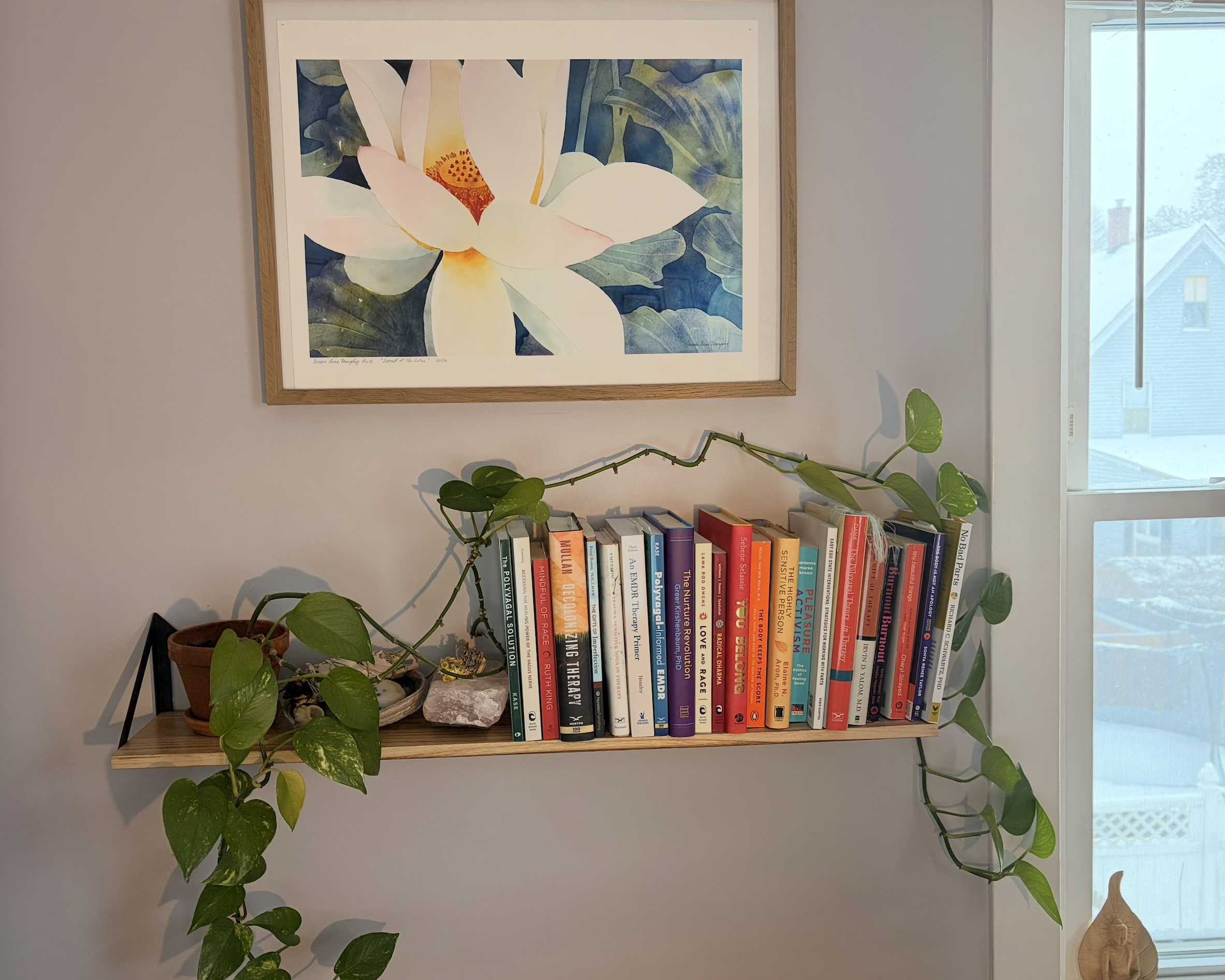 A wooden wall shelf with a potted plant and various books, positioned below a framed painting of a white lotus flower with green leaves, next to a window with a snowy outdoor scene.