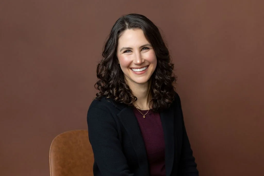 A woman with shoulder-length curly brown hair, smiling, wearing a dark blazer and a burgundy top, sitting on a brown chair against a brown background.