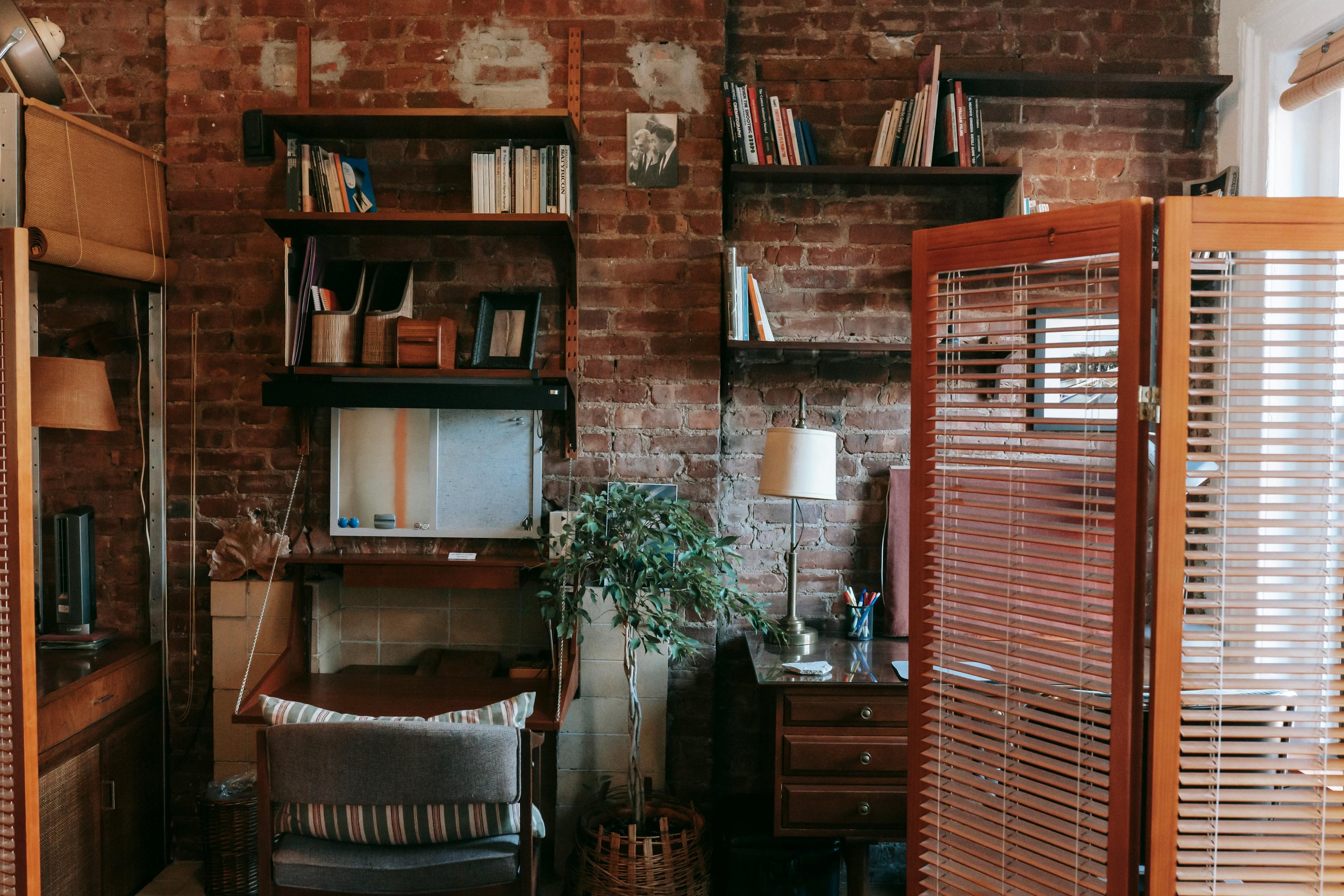 Interior of a room with exposed brick wall, bookshelves, a window, a floor lamp, a small desk with a plant, and furniture including a chair and a room divider.