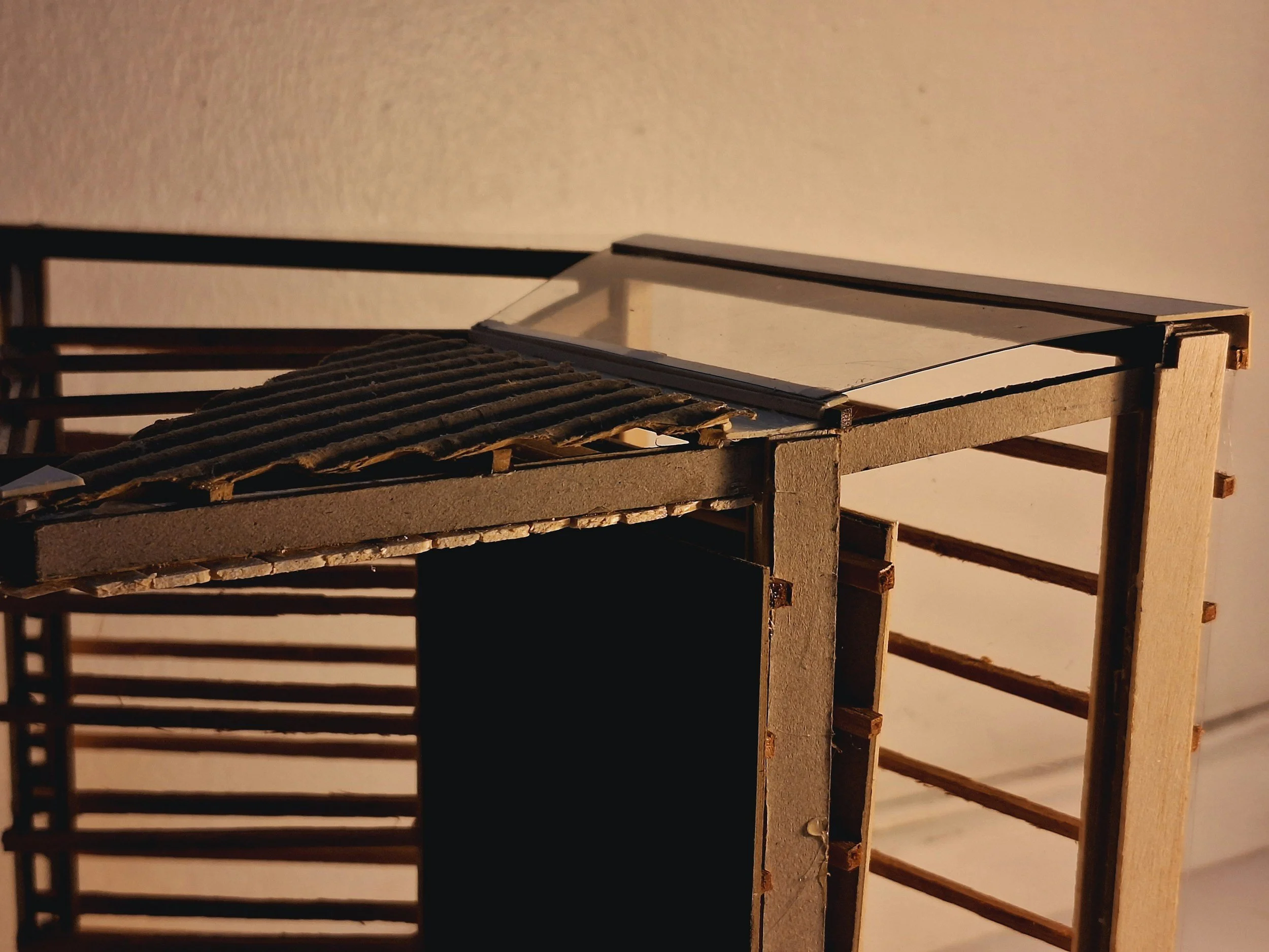 Close-up of a small architectural model or miniature building made from wood, with a glass window and a textured roof section, set against a plain background.