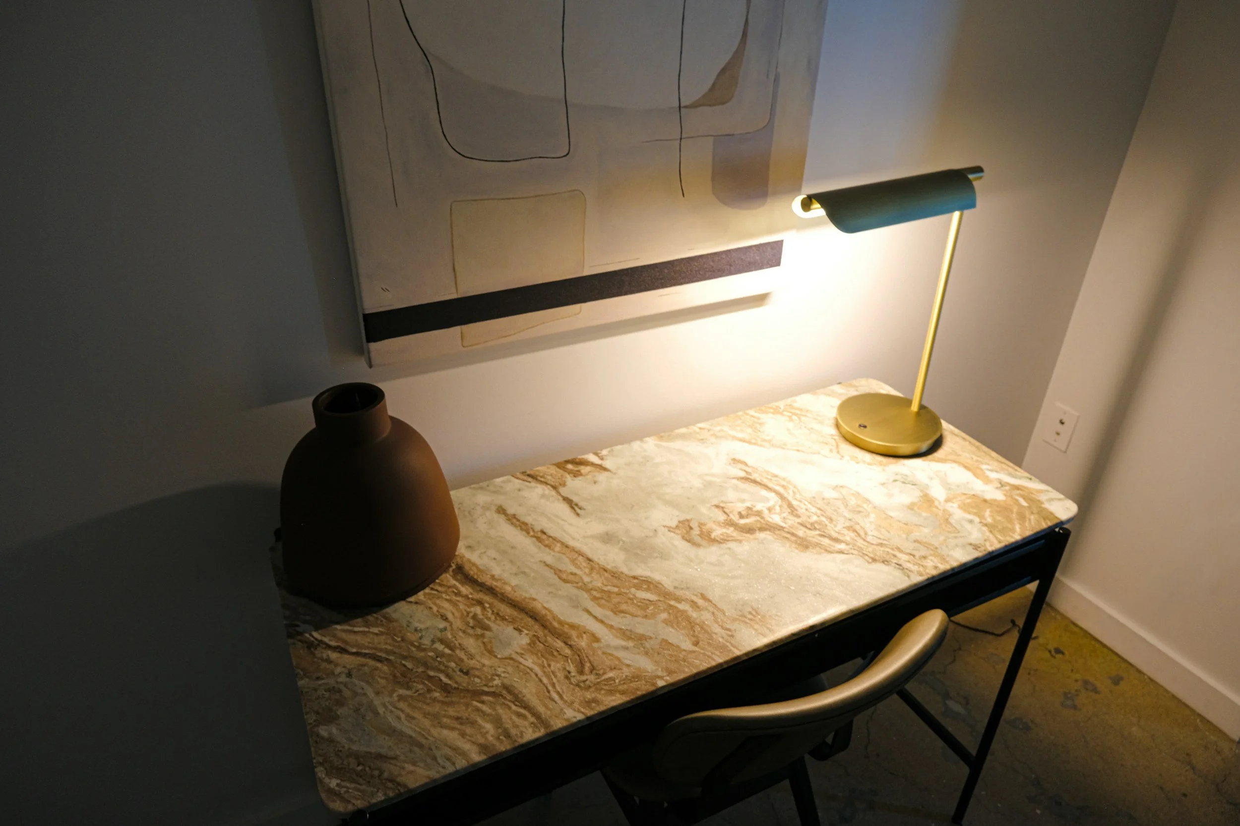 A marble top desk with a brown vase, a gold desk lamp, a chair, and a painting on the wall above it.