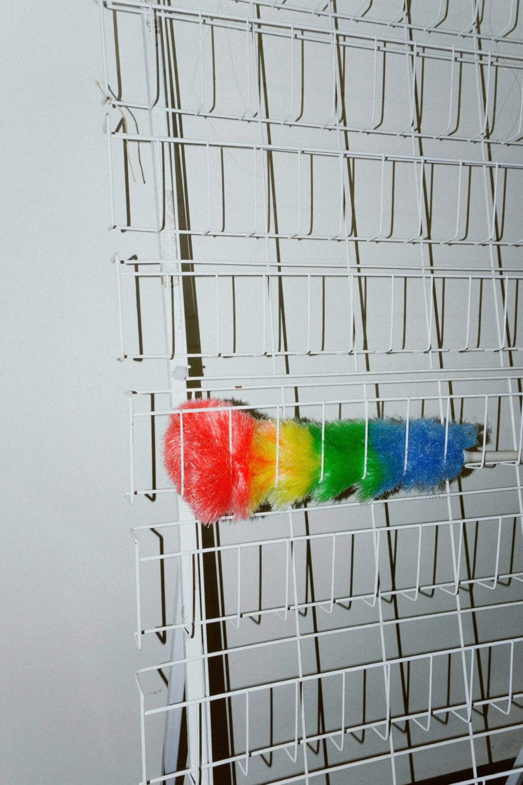 Colorful feather duster with red, yellow, green, and blue feathers hanging on a white wire rack.