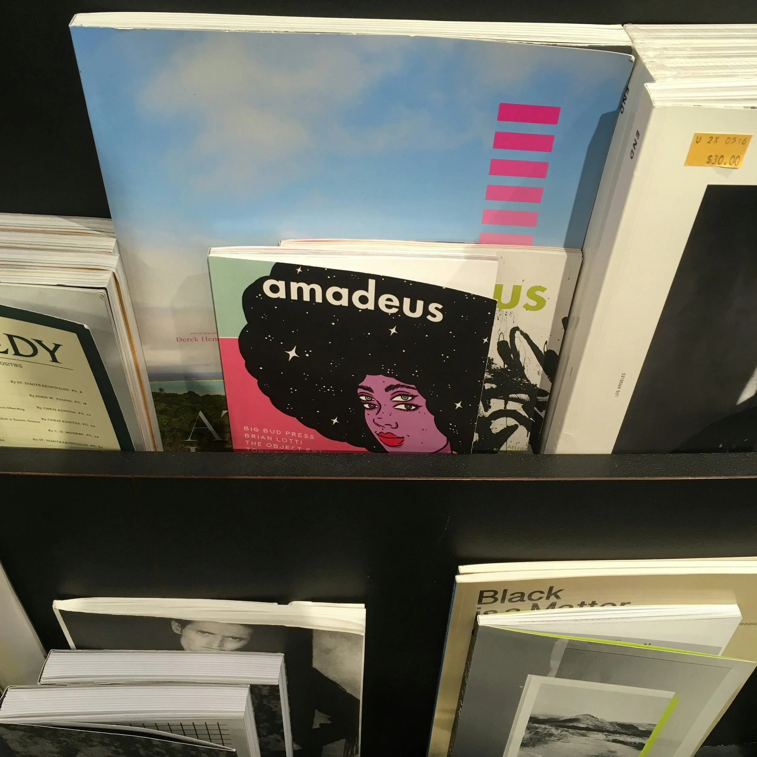A display shelf holding various magazines and art books, with a prominent magazine titled 'Amadeus' featuring an illustrated woman with an afro hairstyle, set against a black starry background.