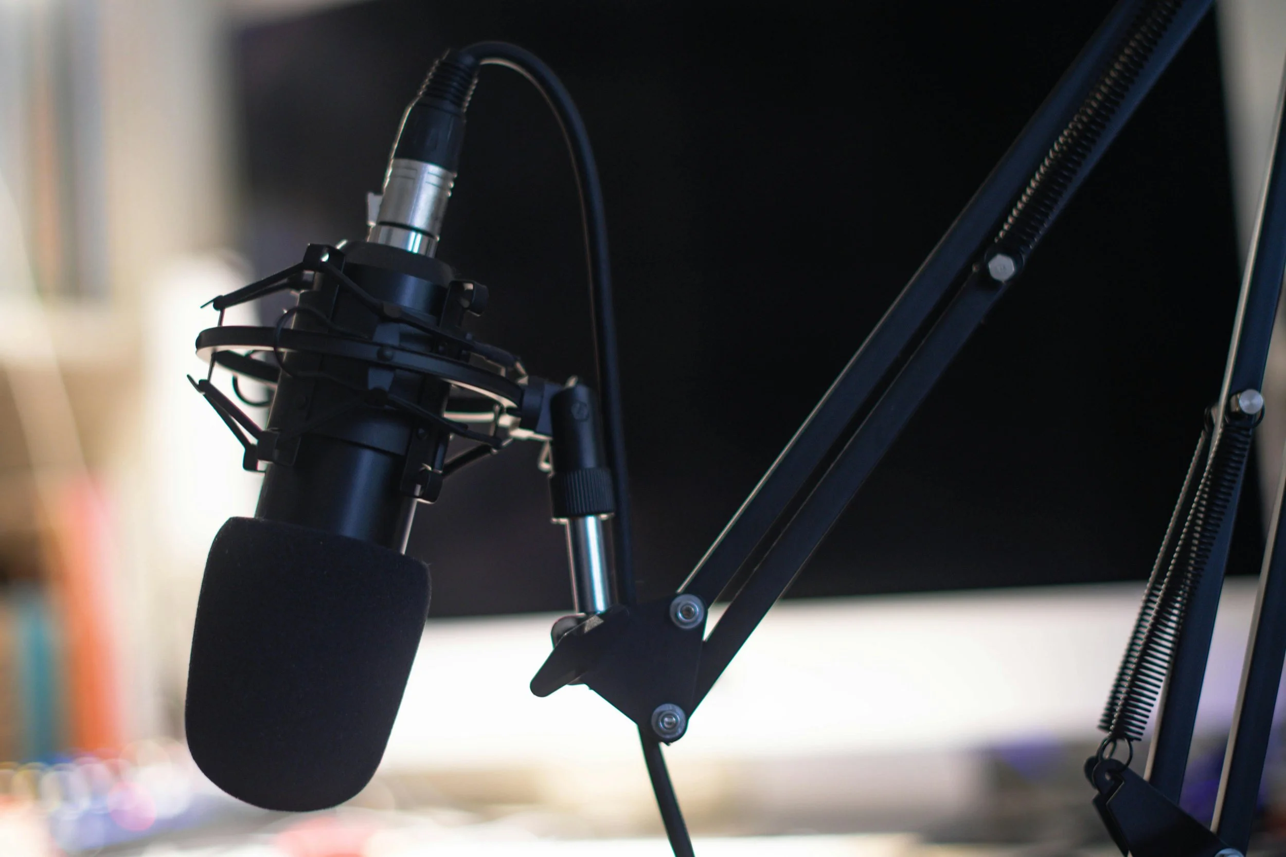 Close-up of a professional microphone mounted on an adjustable arm with a foam windscreen, set up in a recording studio or podcasting setup.
