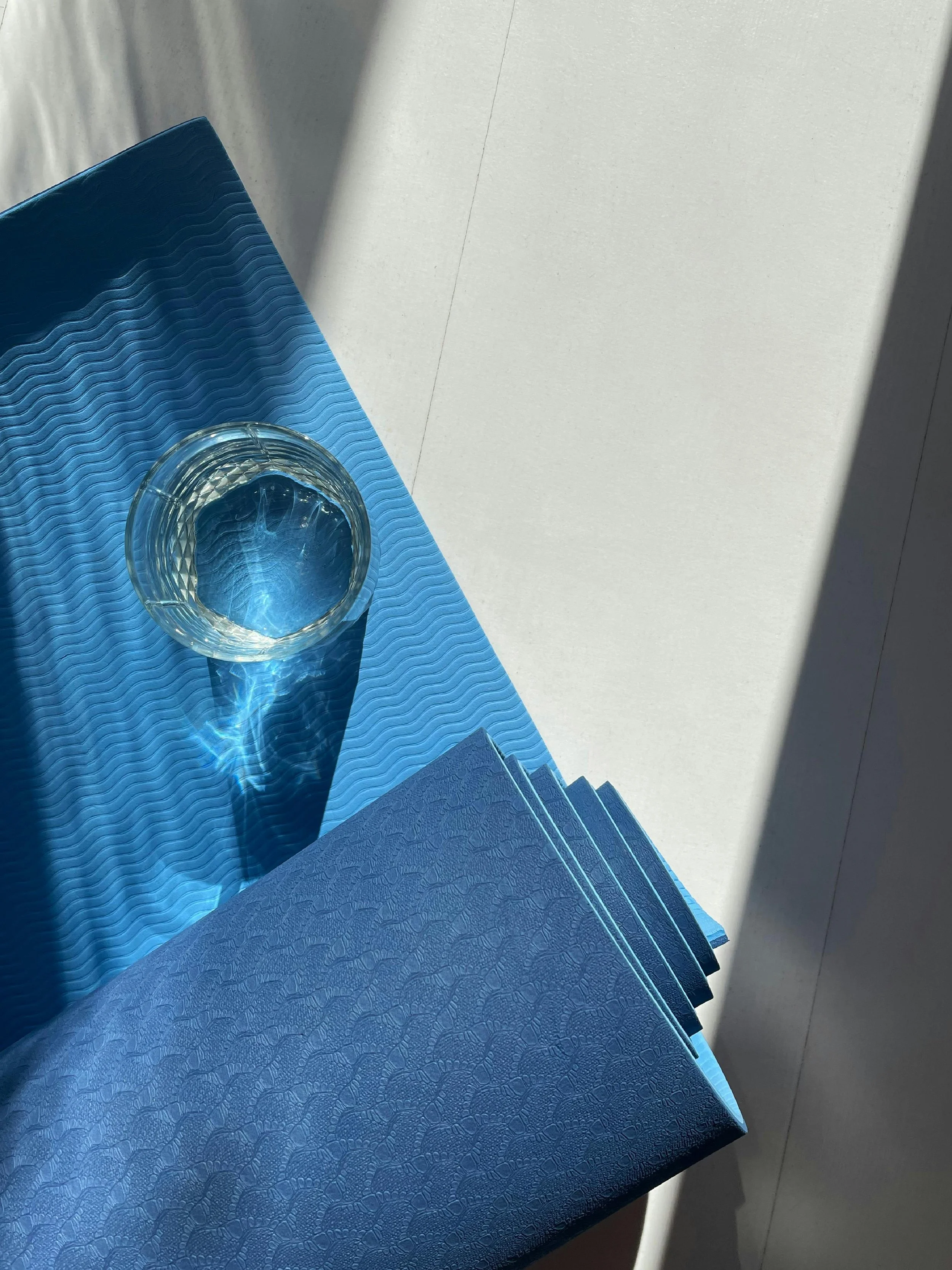 A glass of water placed on a textured blue surface with a pattern, next to a dark blue textured cloth or paper, in sunlight creating shadows and reflections.