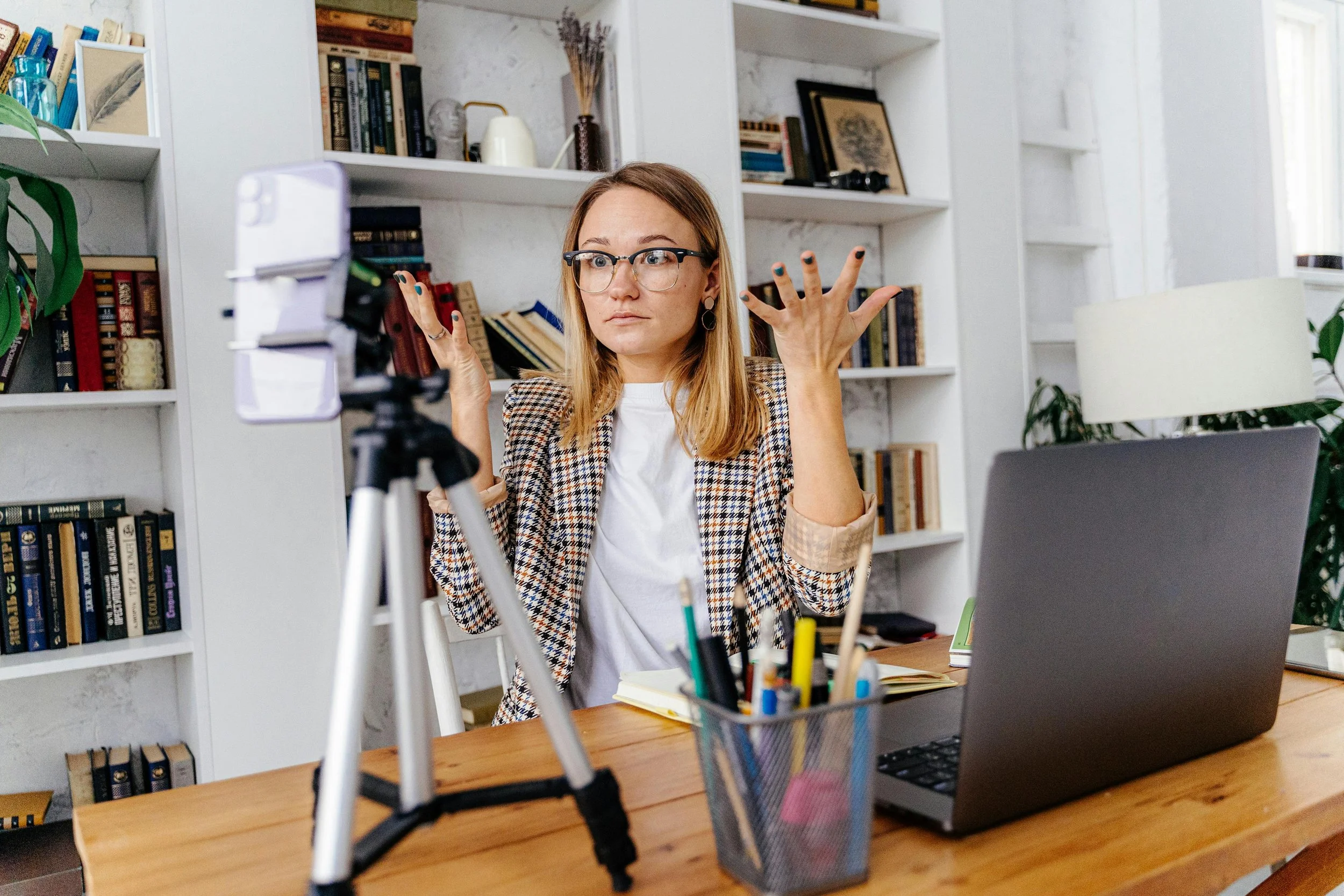 A woman with glasses making expressive gestures in front of a camera on a tripod, in an office setting with bookshelves, a laptop, and writing supplies on the desk.