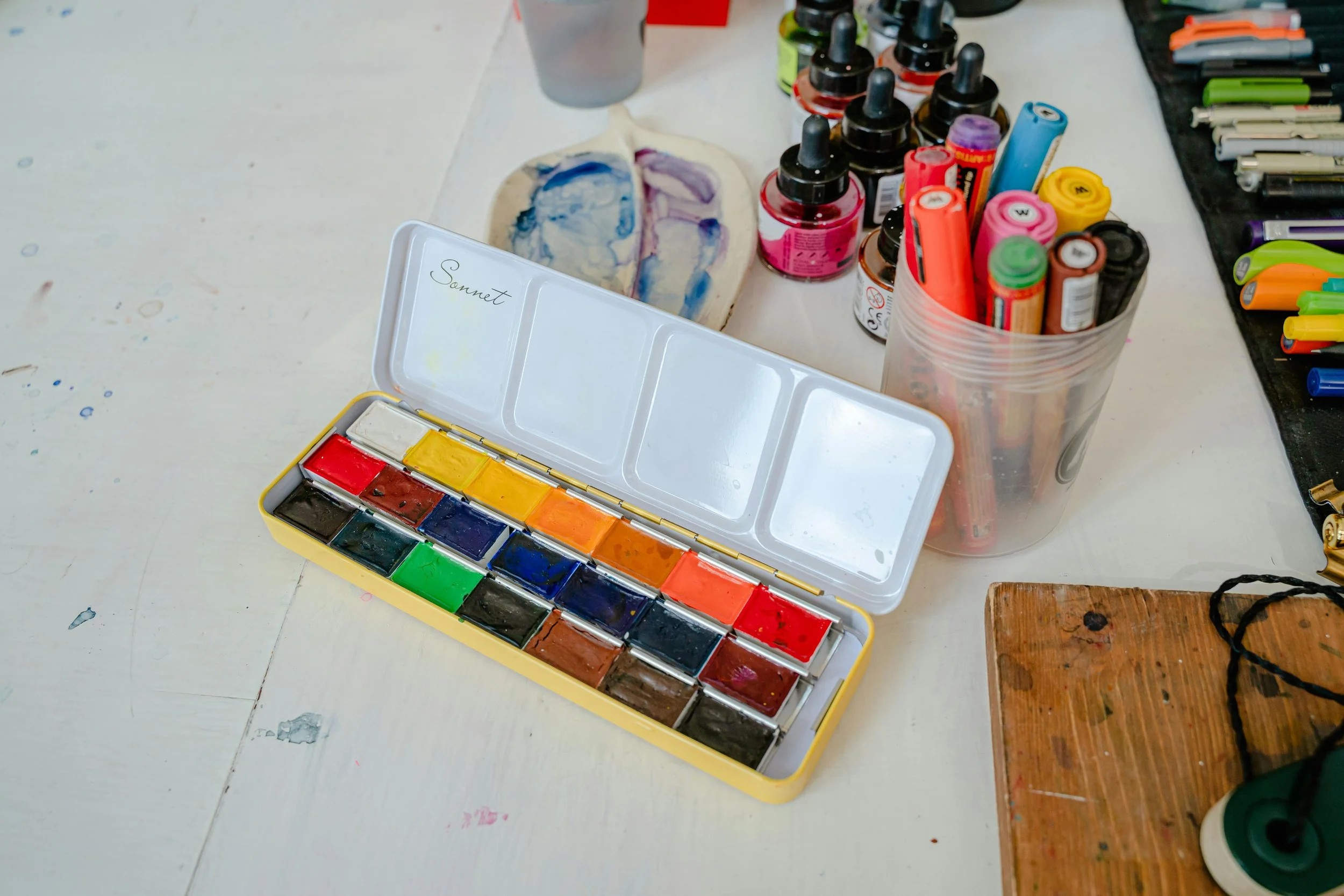 Paint palette with watercolor paints, bottles of ink, markers, and art supplies on a white table.