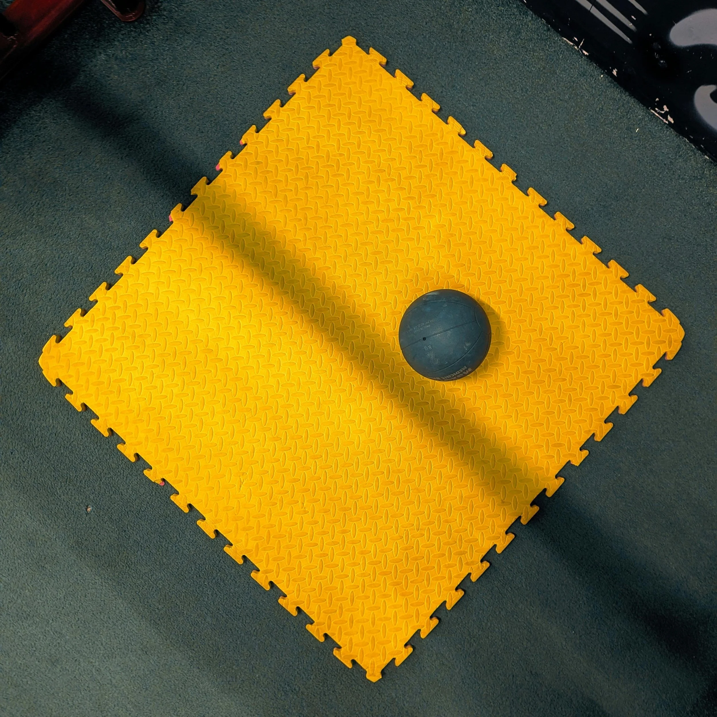 A black medicine ball resting on a yellow interlocking foam exercise mat in a gym or fitness area.