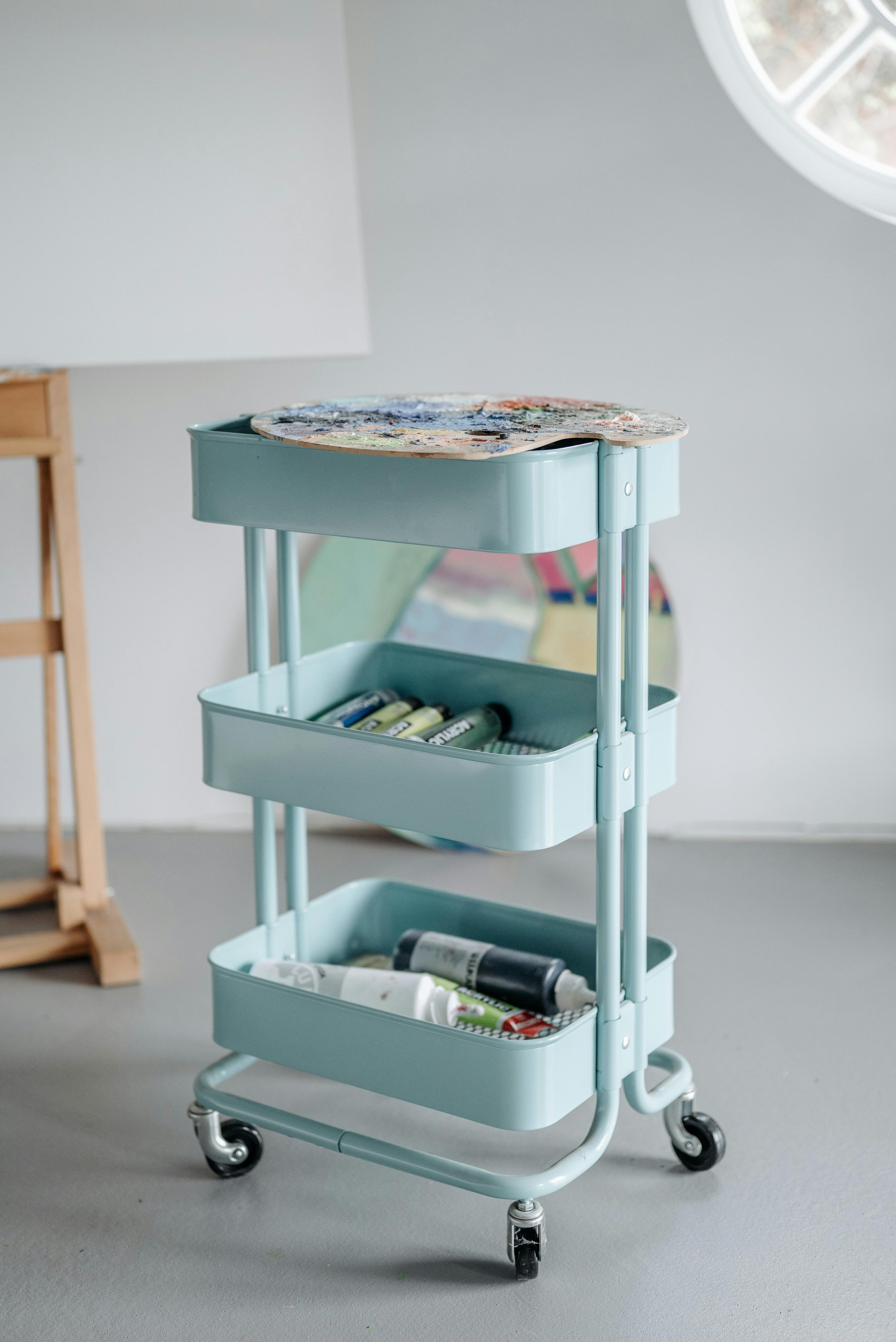 A light blue rolling cart with three shelves holding various art supplies, including paint tubes, markers, and paint palettes, in an art studio with a gray floor and a window.
