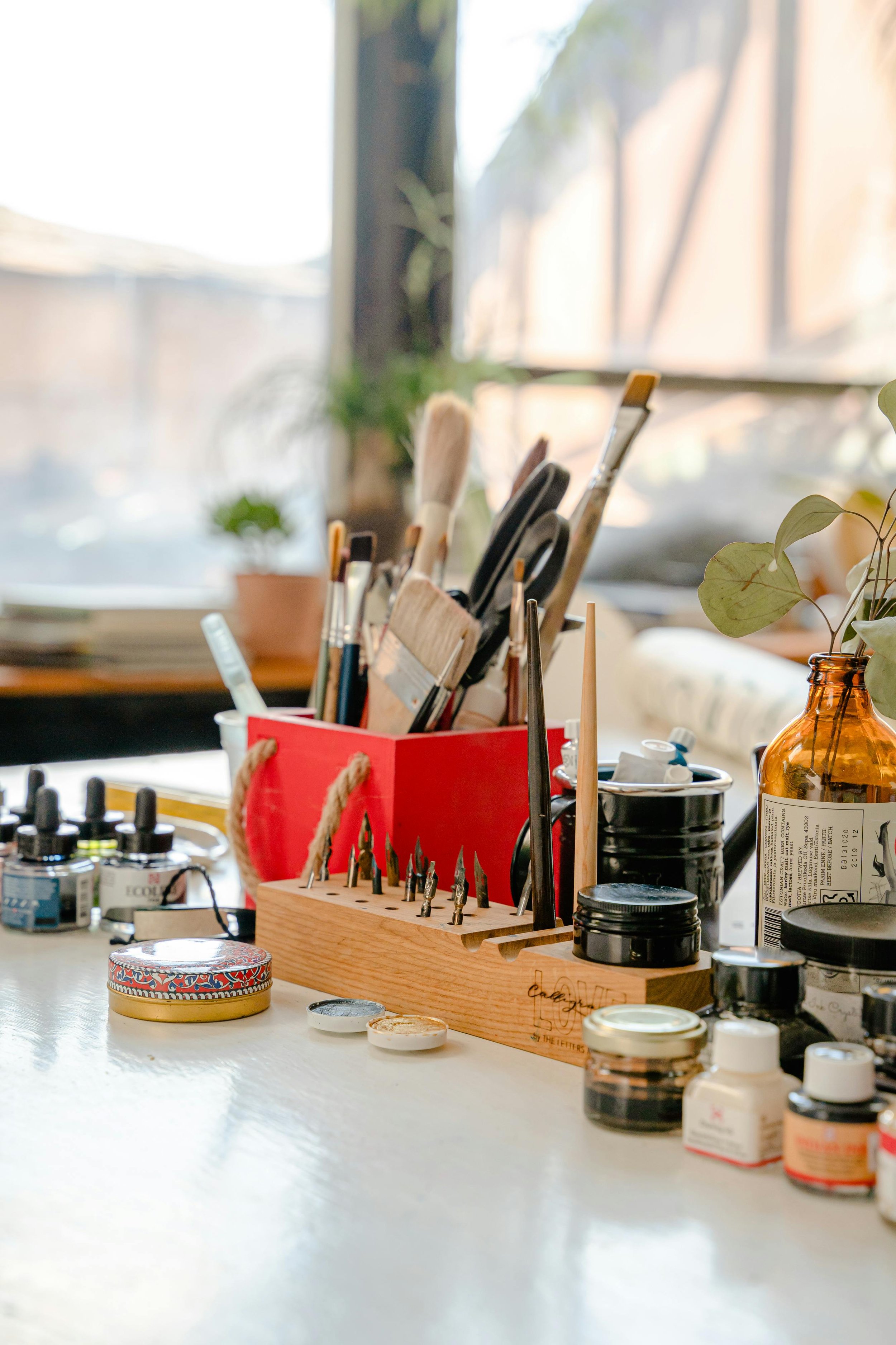 Art supplies, including paintbrushes, pens, and ink bottles, organized on a table near a window with natural light.