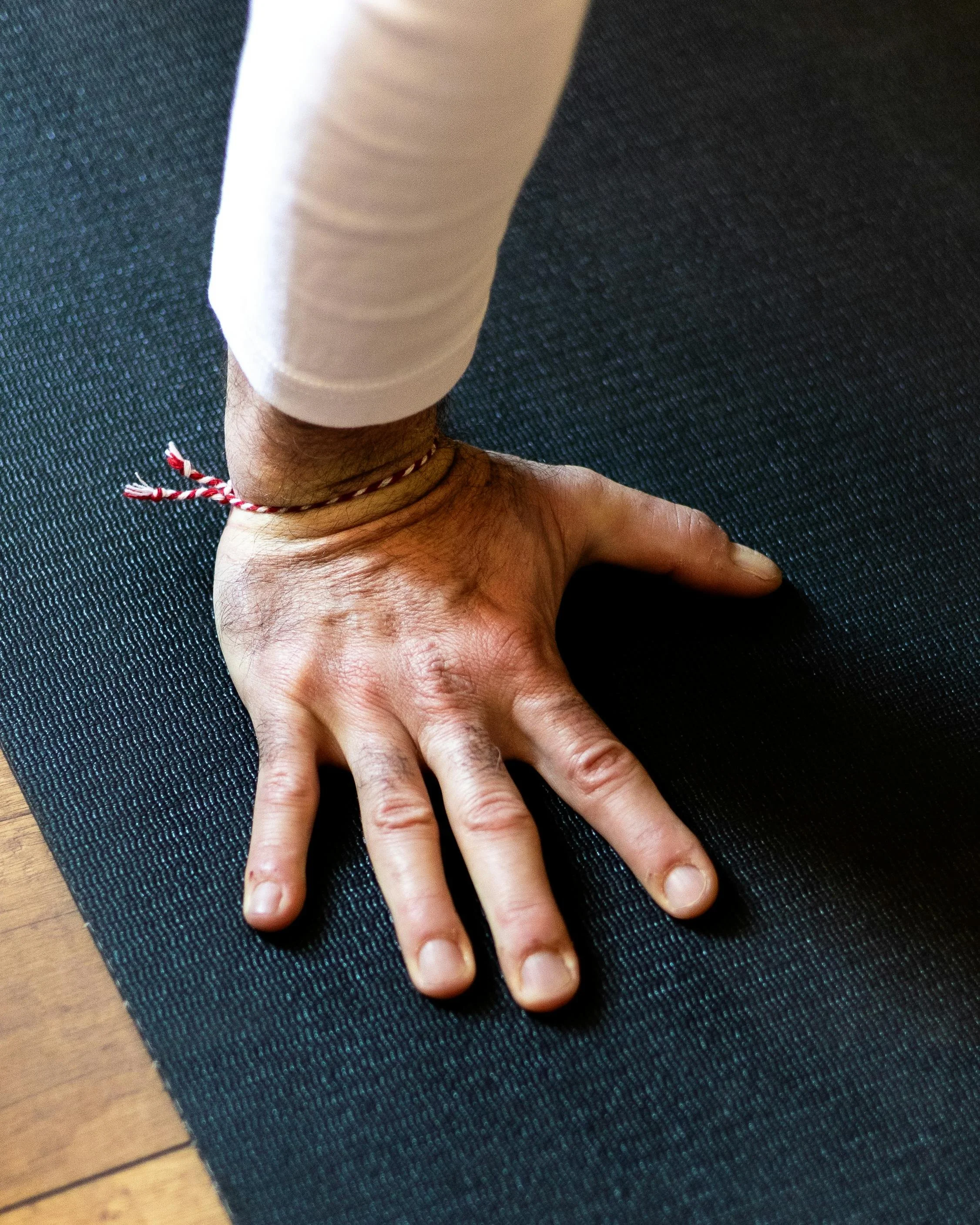 A person presses their hand flat on a textured black yoga mat, wearing a white sleeve and a red-and-white string bracelet.