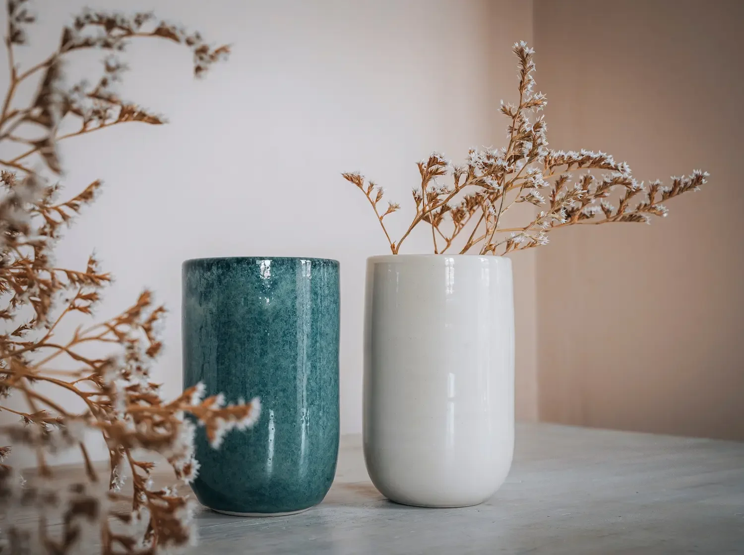 Deux vases en céramique, un bleu et un blanc, avec des branches de fleurs séchées, placés sur une table en bois.