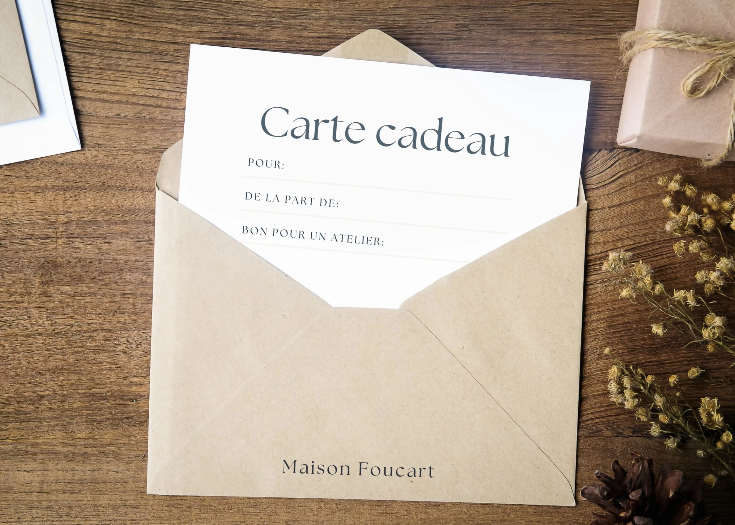 Une carte cadeau dans une enveloppe en papier kraft posé sur une table en bois, accompagnée de petits paquets emballés, de plantes séchées et de photos.