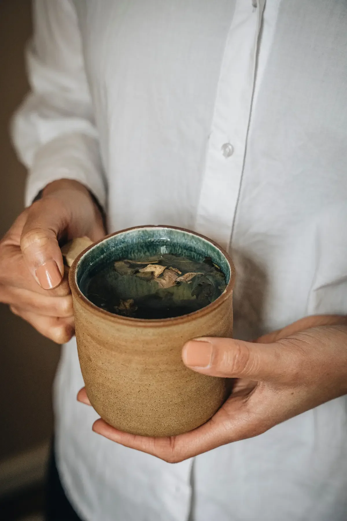 Une personne tient une tasse en céramique contenant une infusion d'herbes ou un thé, avec une chemise blanche à manches longues.