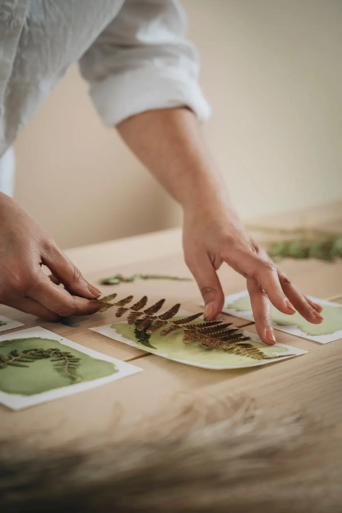 Une personne manipule des feuilles de papier avec des empreintes ou des impressions de feuilles de fougère, posées sur une table en bois.