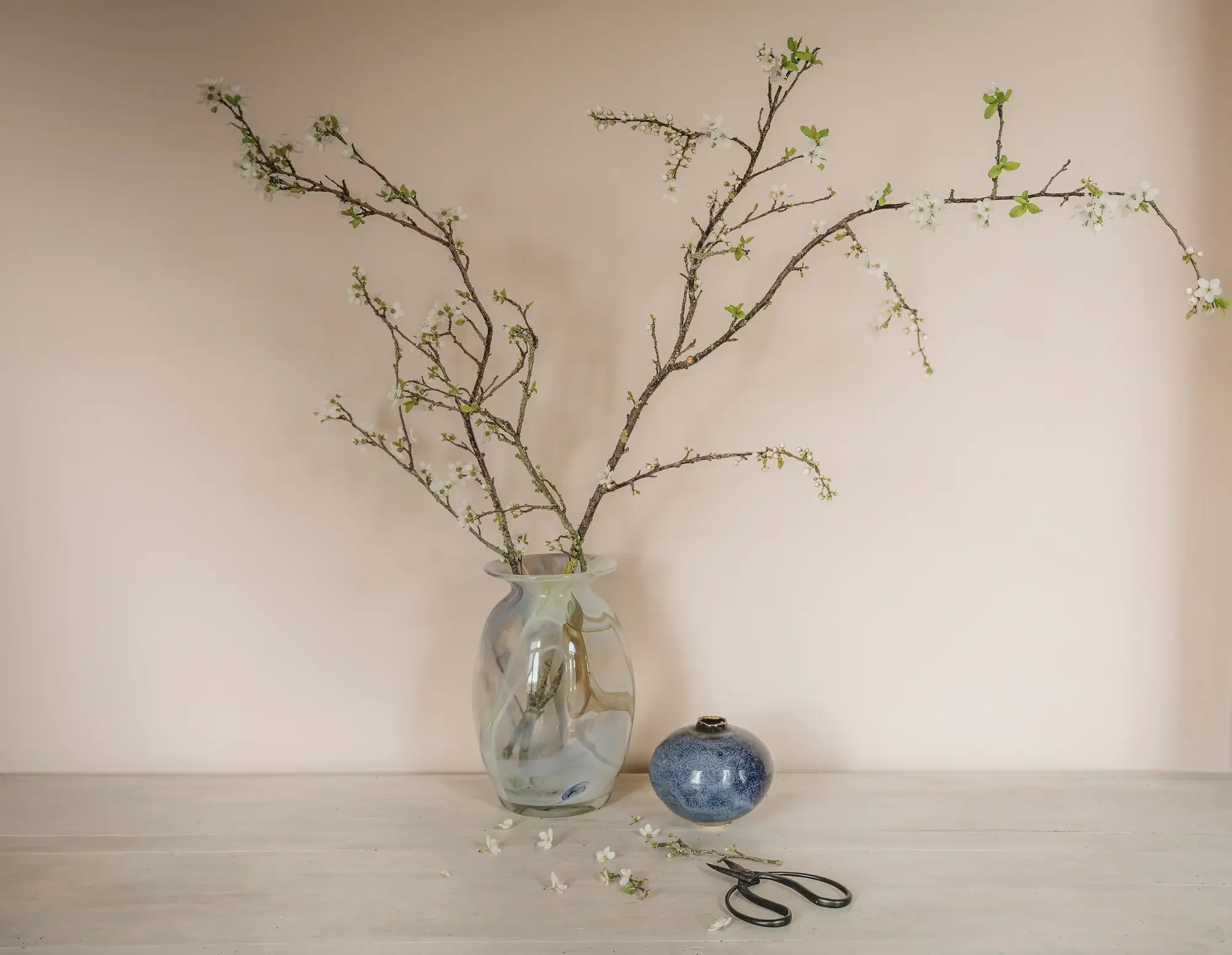 Branche florale dans un vase en verre et un petit vase en céramique sur une table en bois, avec des ciseaux.